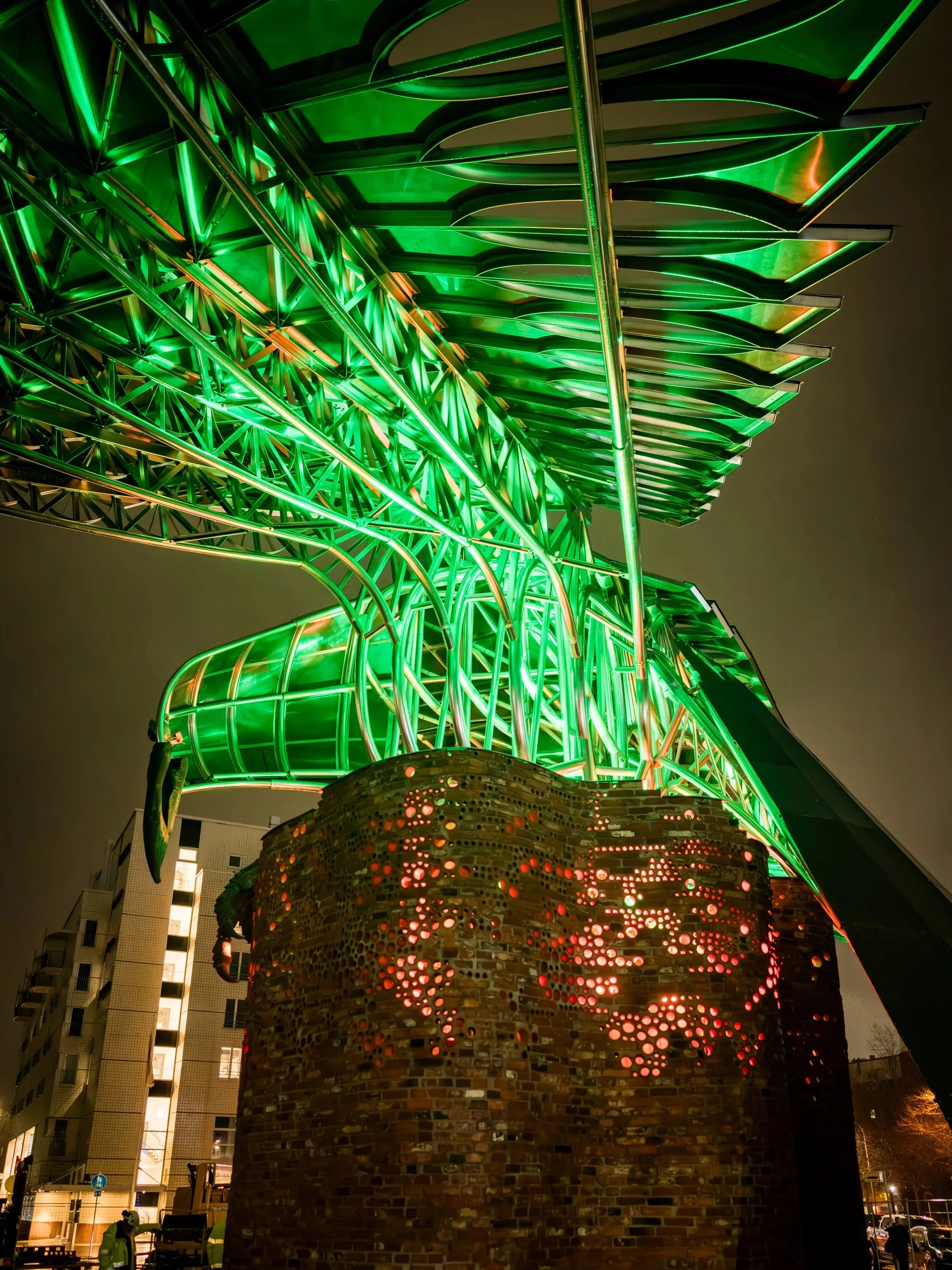 Yöllisessä ulkotilassa kirkkaasti valaistu korppiveistos seisoo tiilijalustalla, toinen siipi levittäytyneenä kohti katsojaa ja valaistus on vihreä.