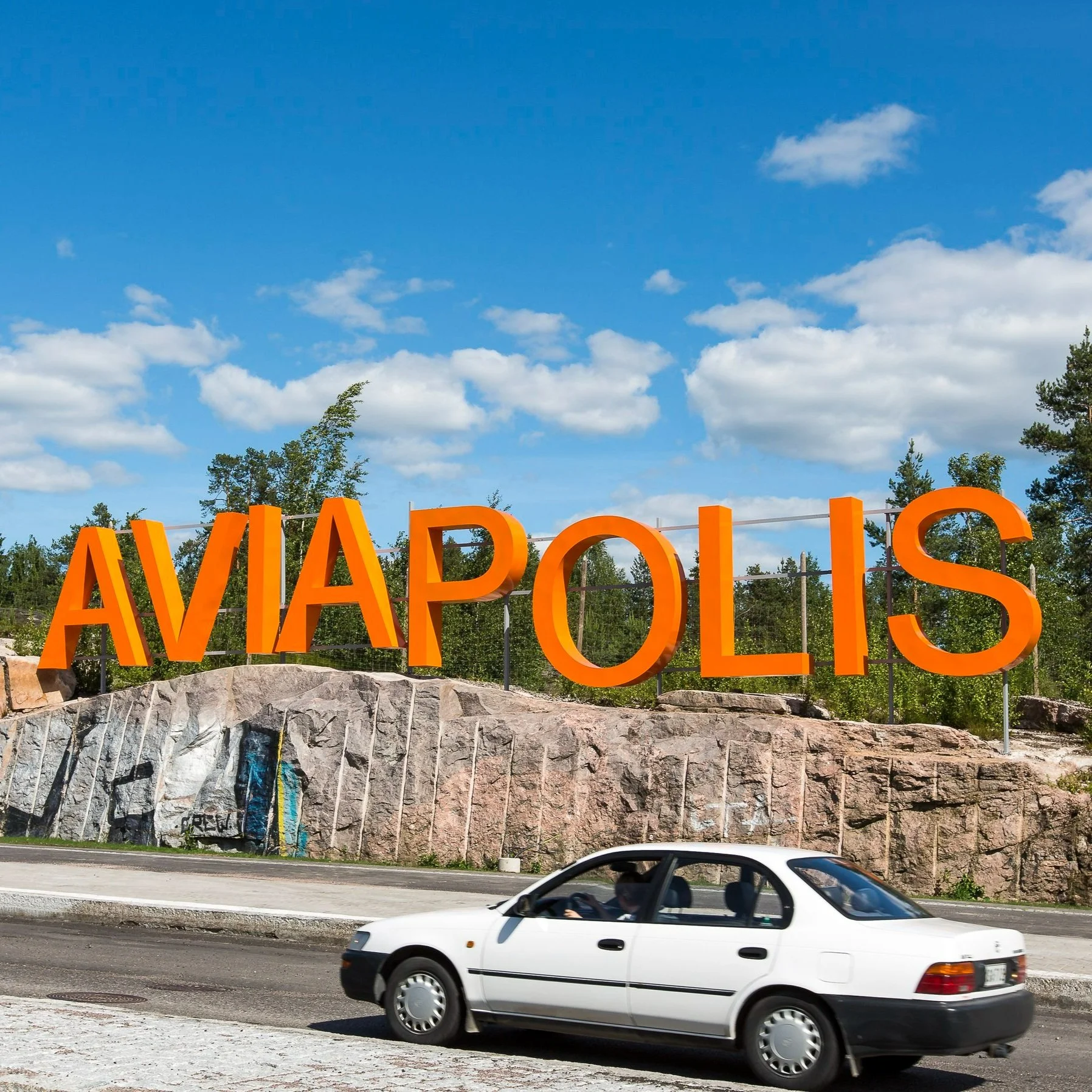 Valkoinen henkilöauto ajaa kesäisellä tiellä, taustalla näkyy kallioseinämä, jonka päällä on suuri oranssi Aviapolis-kyltti, havainnekuva.