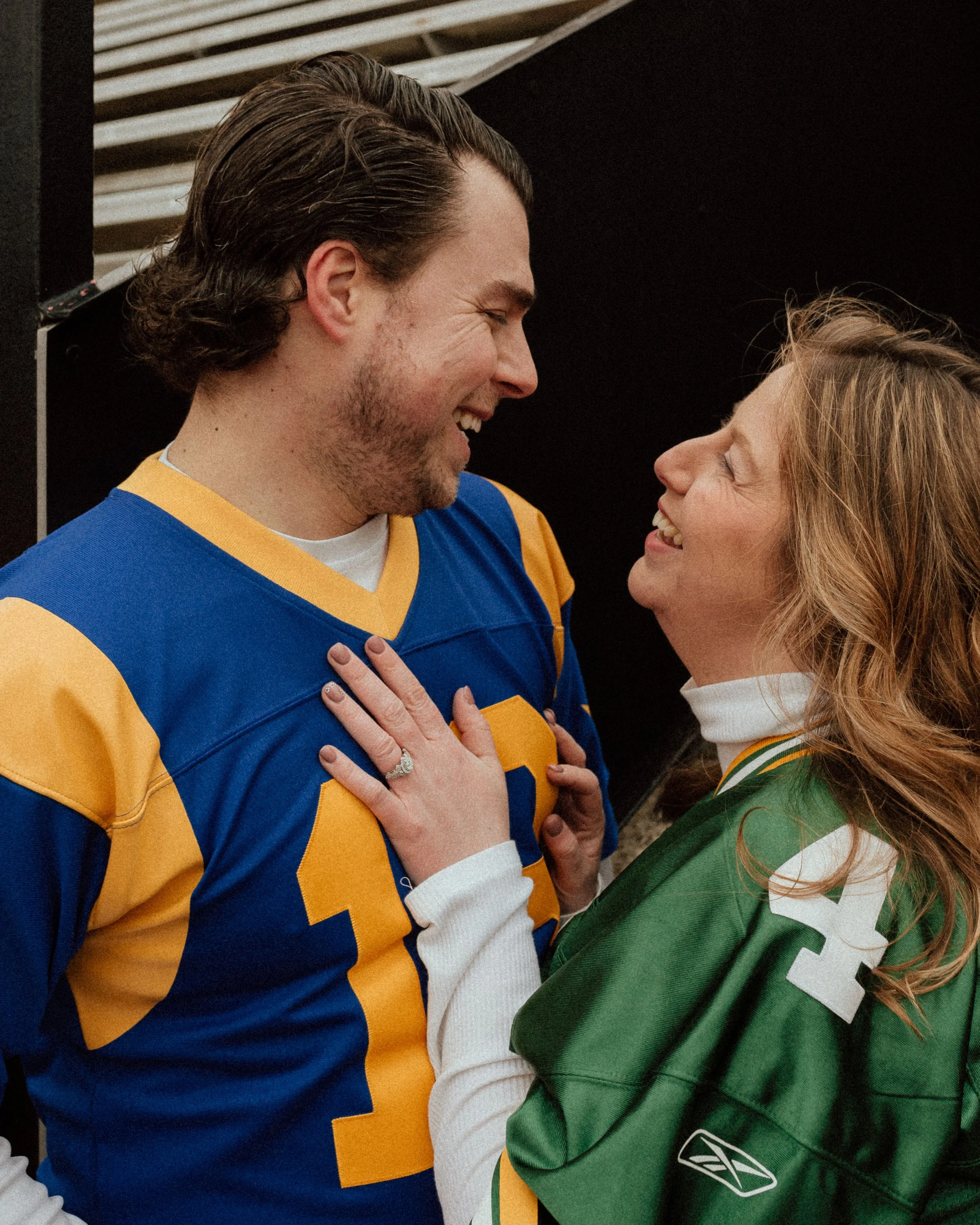 A couple looking at each other laughing while embracing in football jerseys.