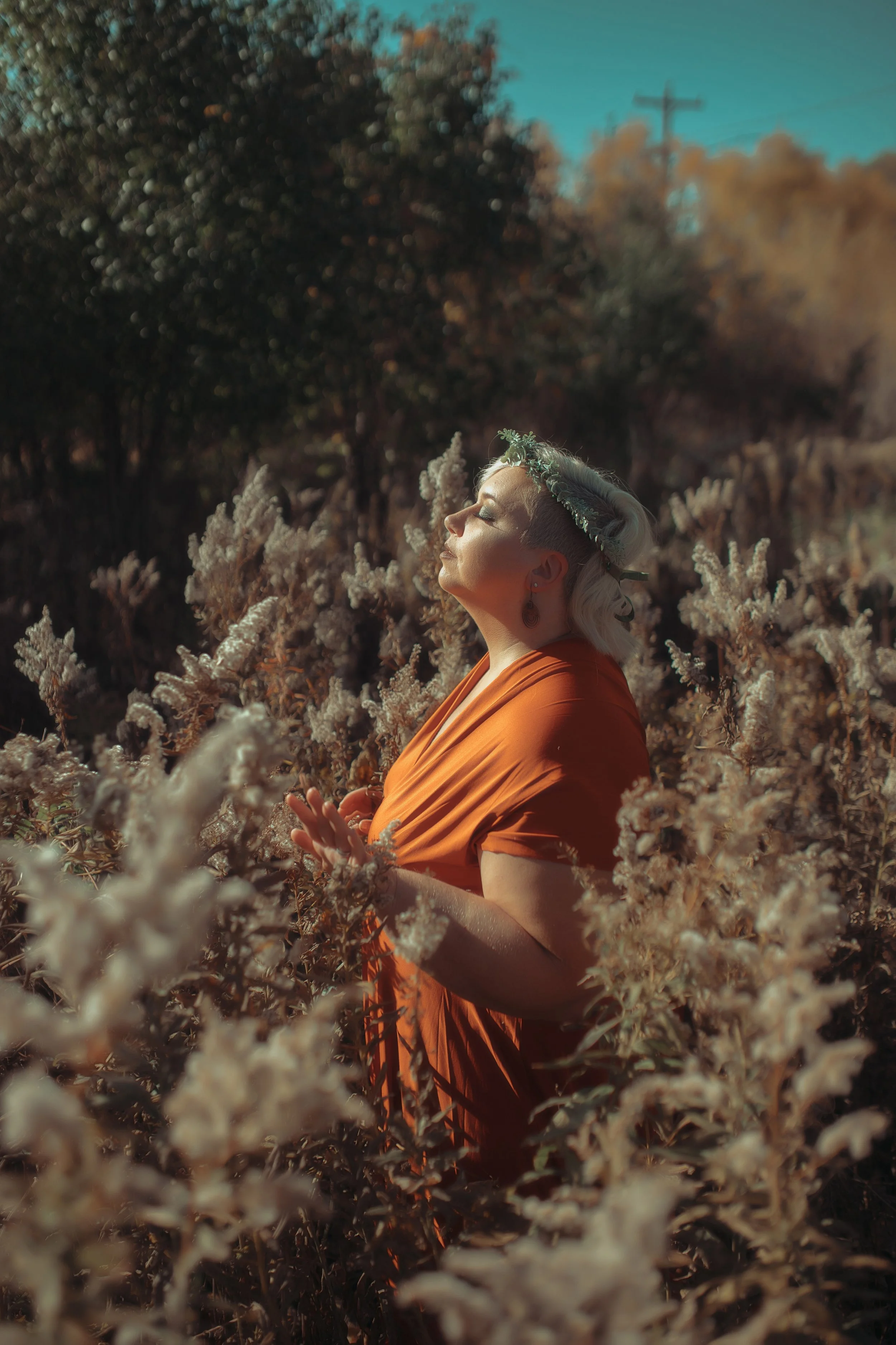 Woman in orange dress with a floral crown standing among white flowering plants in a field with trees and power lines in the background, basking in sunlight with eyes closed.
