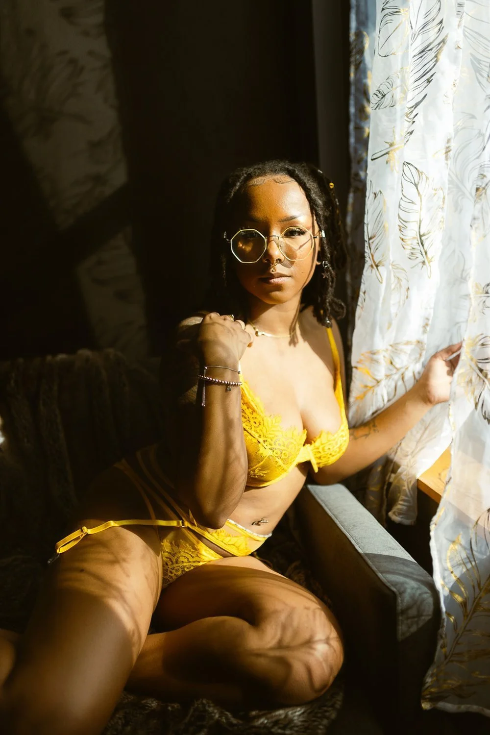 Woman in yellow lingerie sitting on a sofa near a window with patterned curtains, holding the curtain with one hand and looking at the camera.