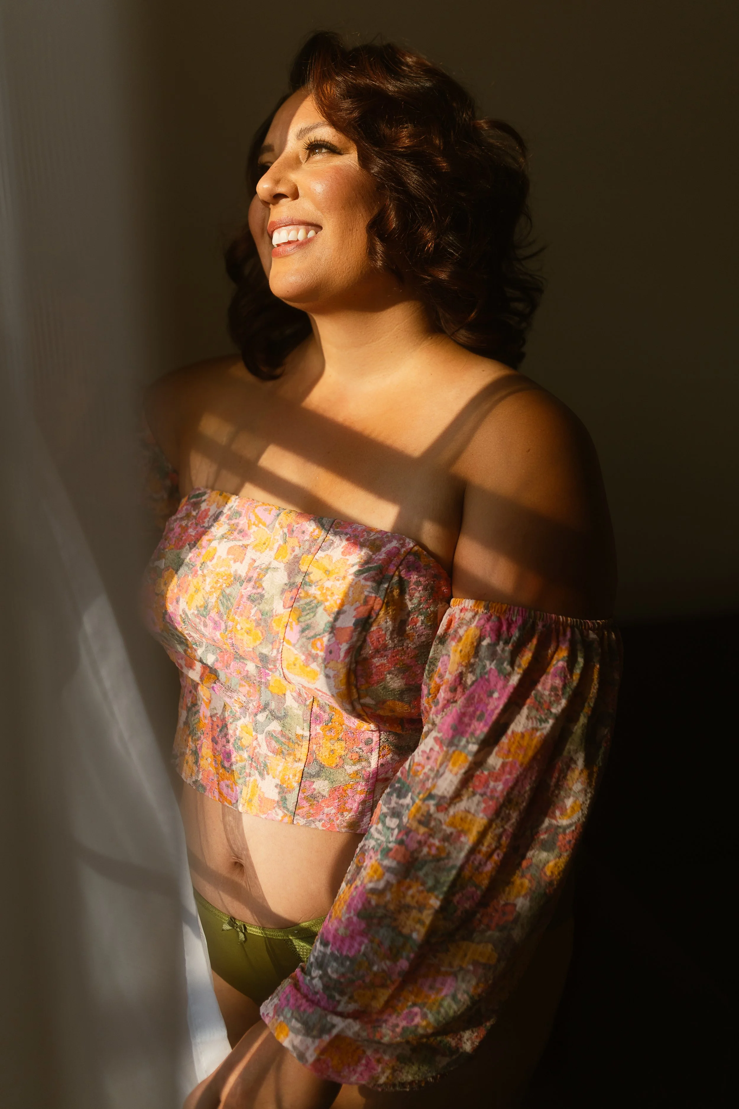 Smiling woman with dark brown curly hair, wearing a floral off-the-shoulder top with puffy sleeves, standing in sunlight with shadows cast across her face and body.