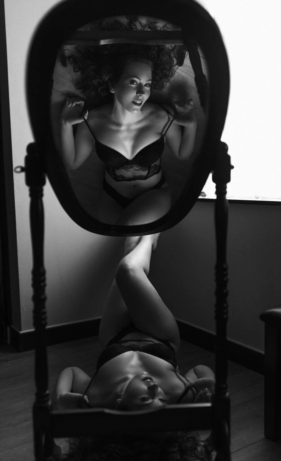 A woman in black lingerie lying on the floor, her reflection visible in a mirror placed vertically in front of her, with her head at the top and legs crossed at the bottom of the mirror.