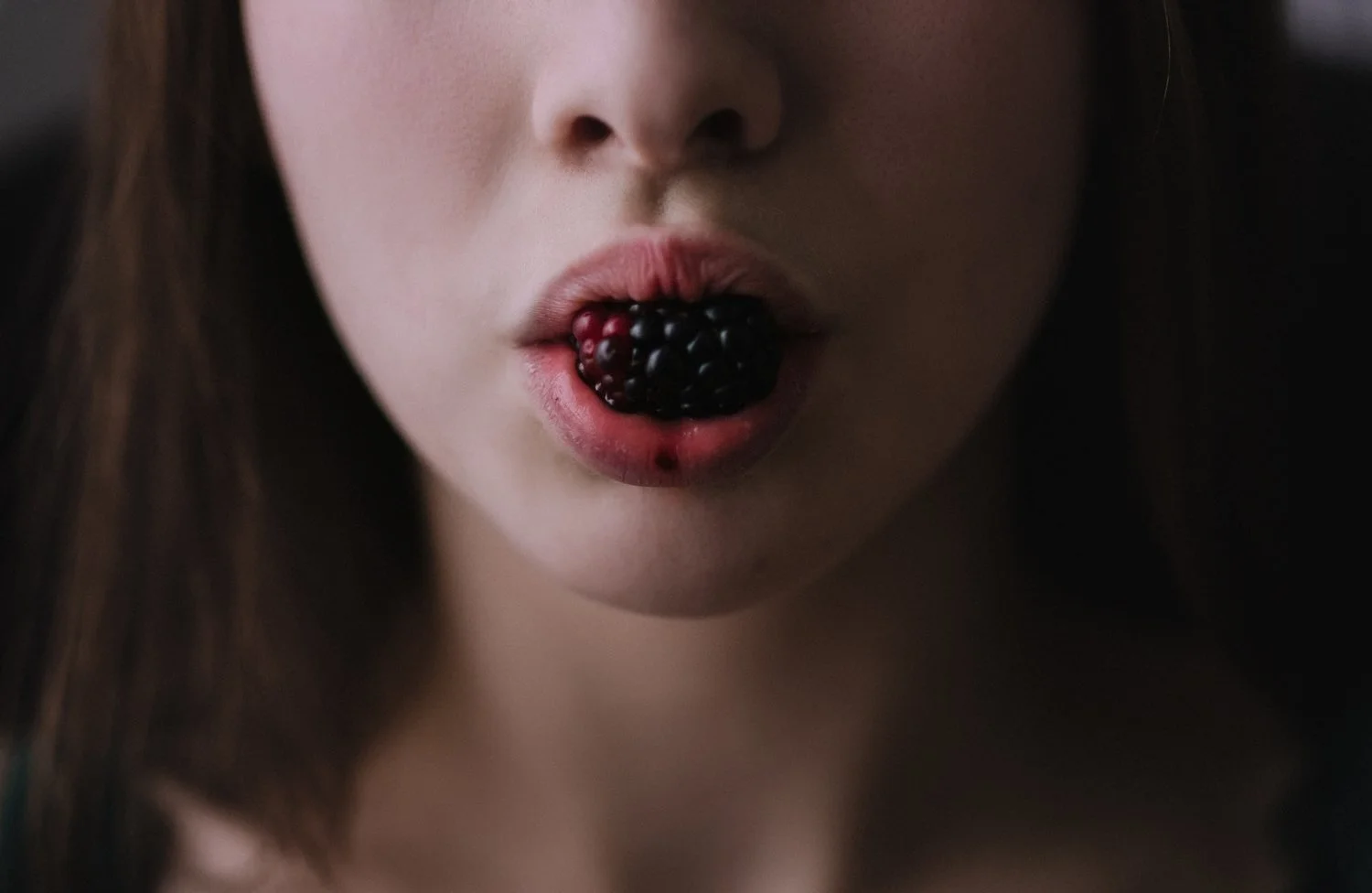 A close-up of a woman's face with her mouth slightly open, holding a cluster of blackberries between her lips, with berry juice on her lips.