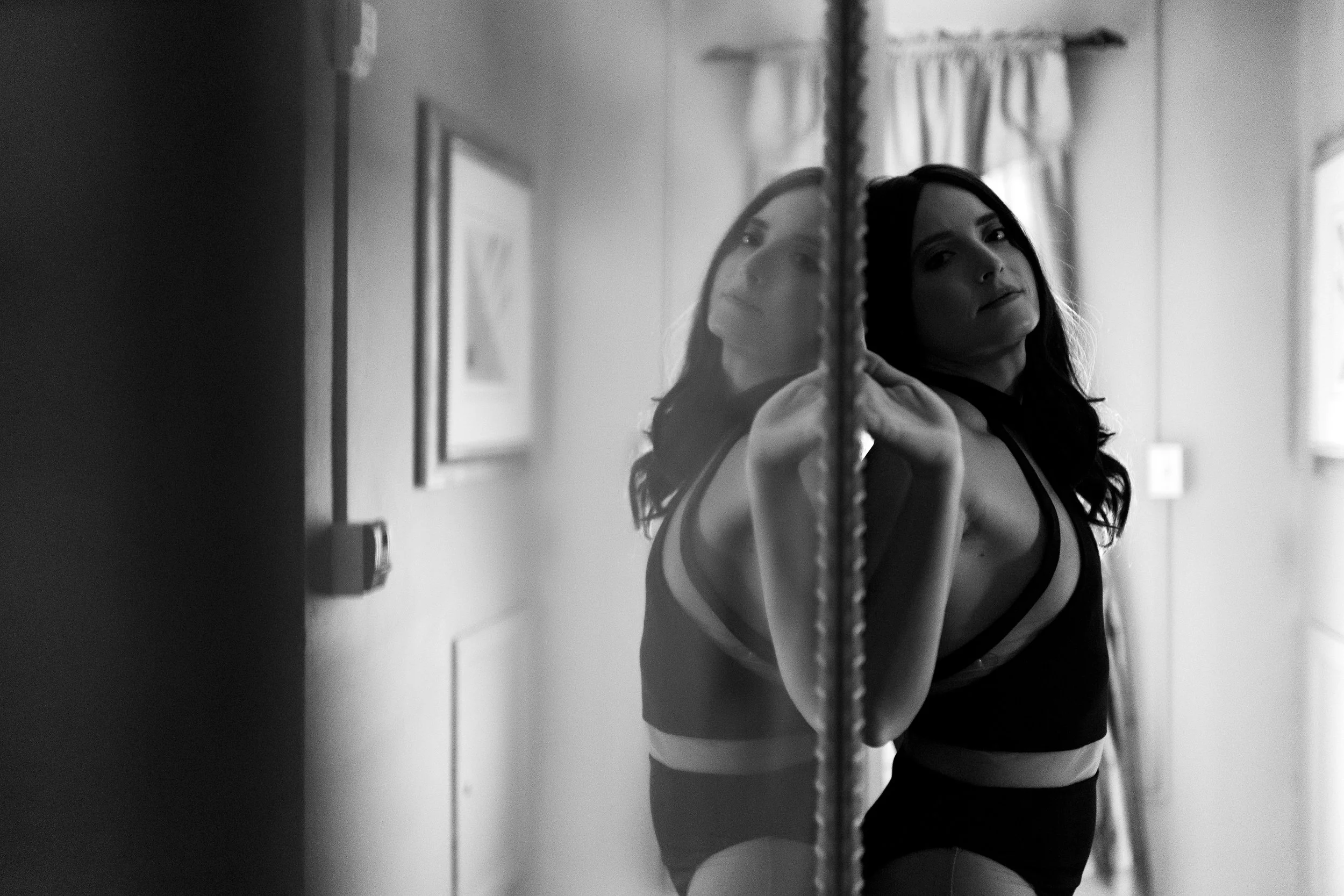 A woman with dark hair looking at a mirror, with her reflection visible. She is wearing a black and white top. The image is in black and white, taken indoors with natural light from a window.