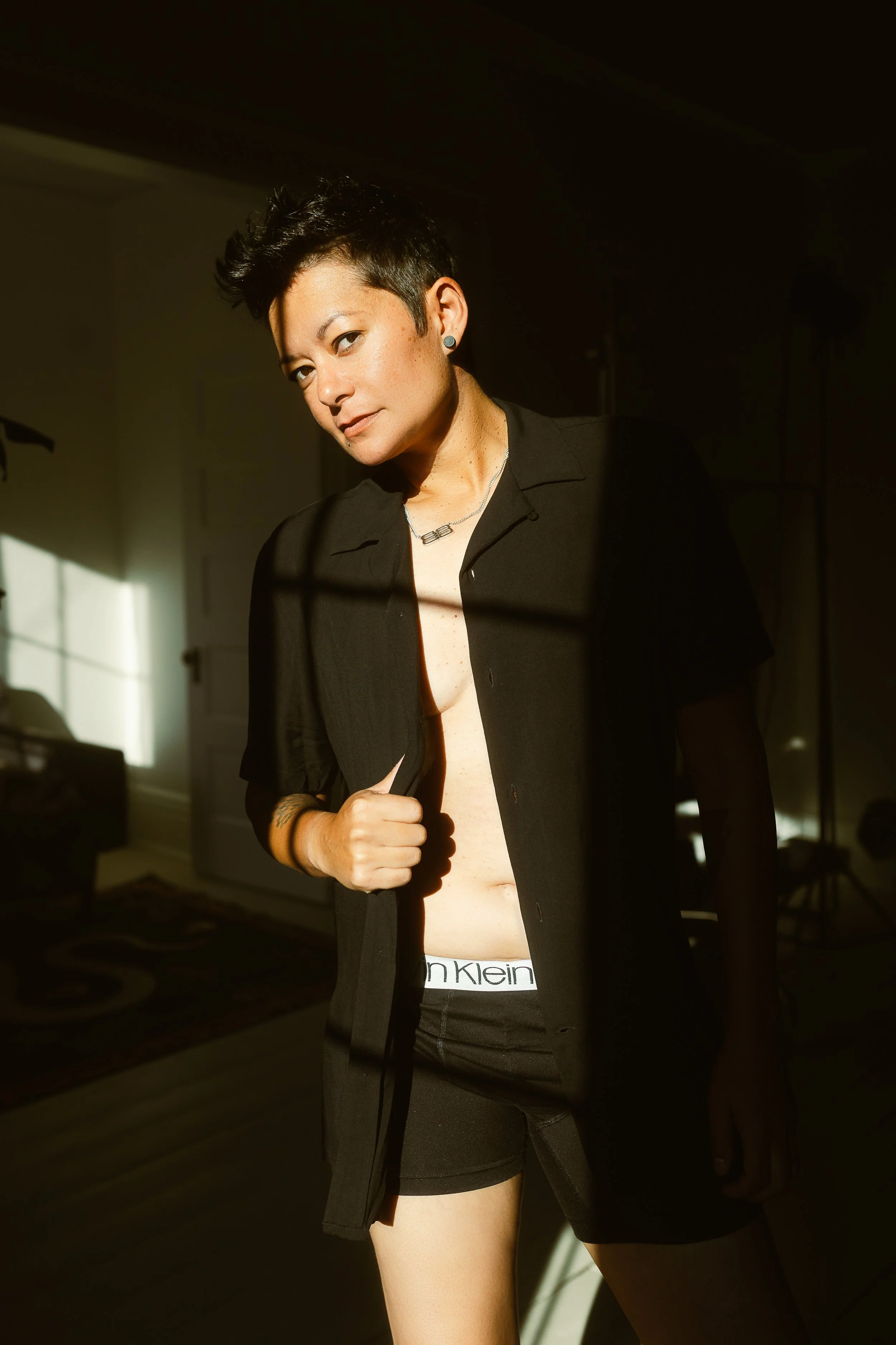 Young woman with short dark hair partially unbuttoning a black shirt in sunlight, inside a room.