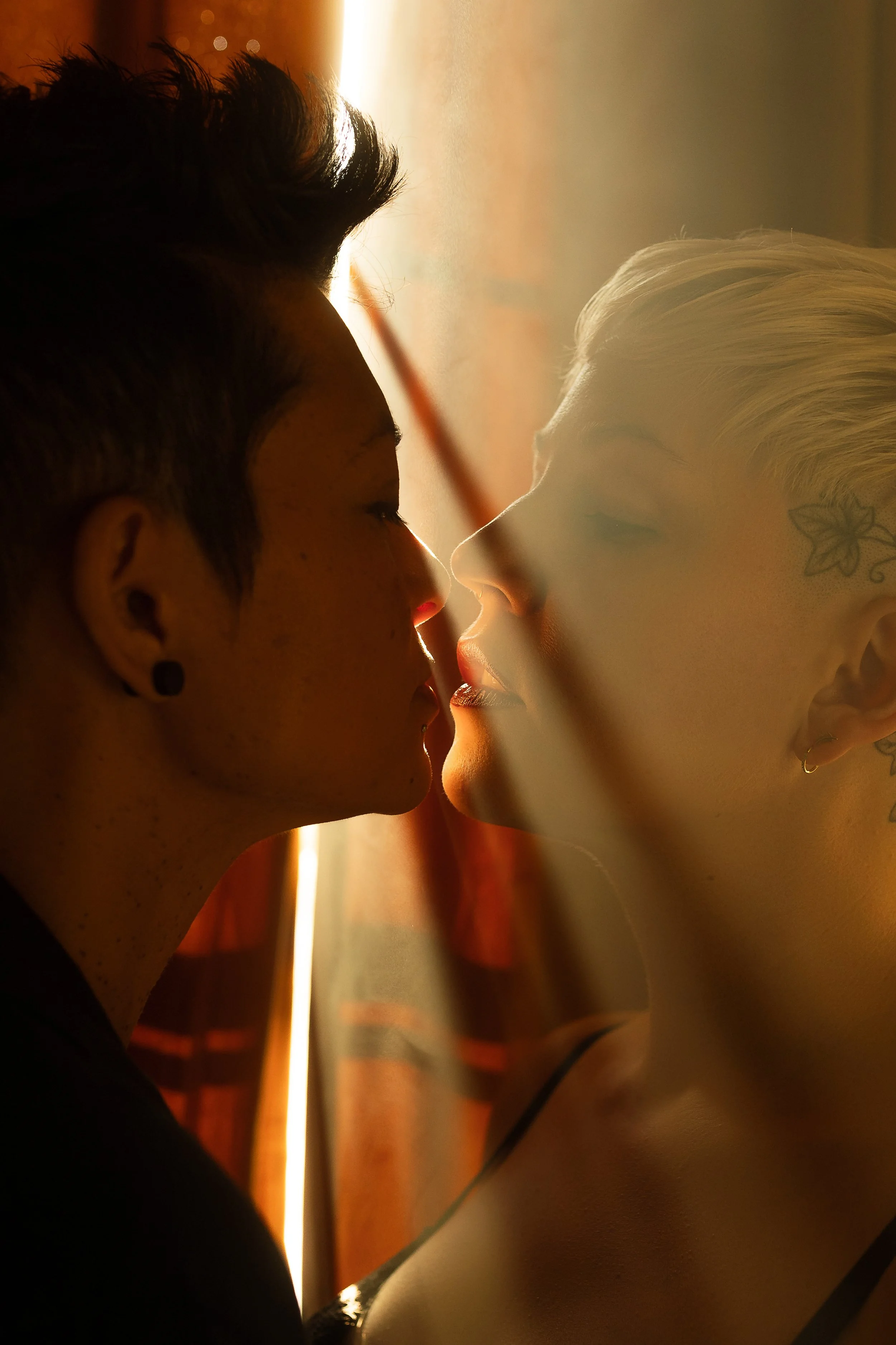 Two people with short hair and tattoos are close to each other, touching noses, with their eyes closed, backlit by warm sunlight.