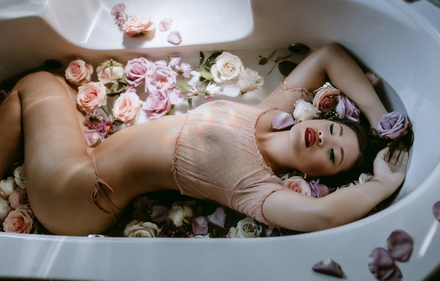 A woman lying in a bathtub filled with rose petals, wearing a pink top and underwear with her eyes closed.