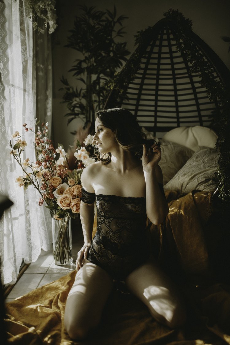 A woman sitting on a bed in front of a window with lace curtains, wearing black lace lingerie, with a large bouquet of flowers nearby, and a decorative headboard behind her.