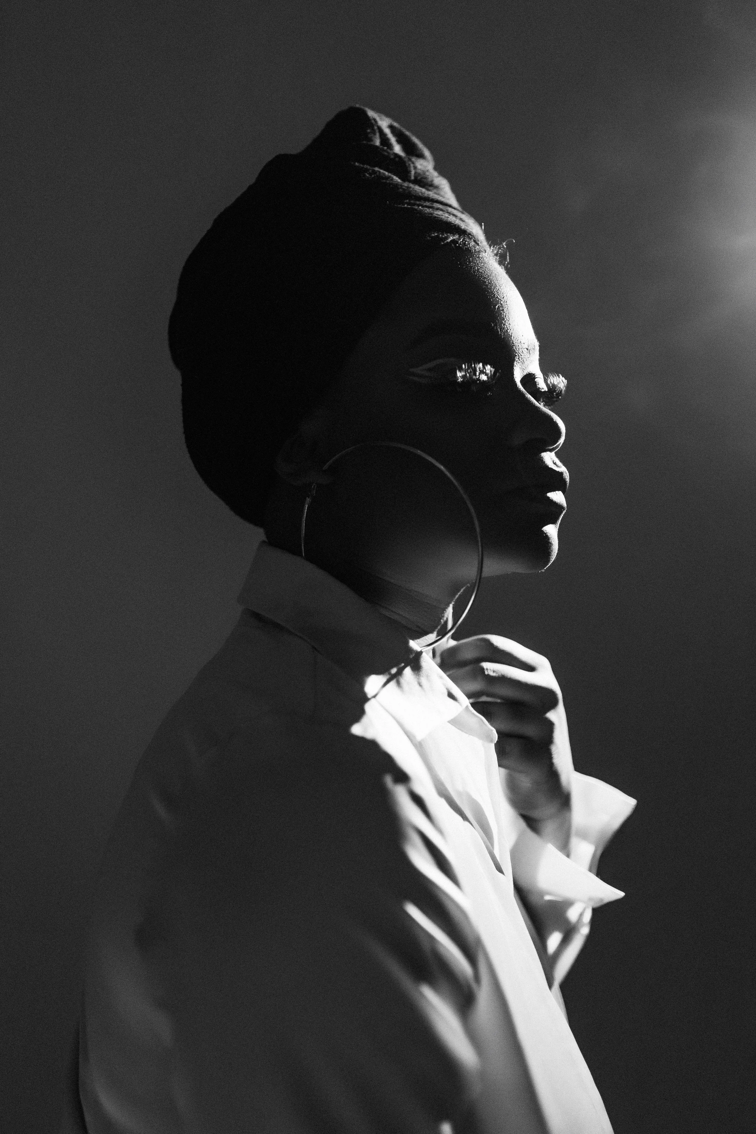 A black and white portrait of a woman in profile, wearing a head wrap and large hoop earrings, with sunlight illuminating her face from the right side.