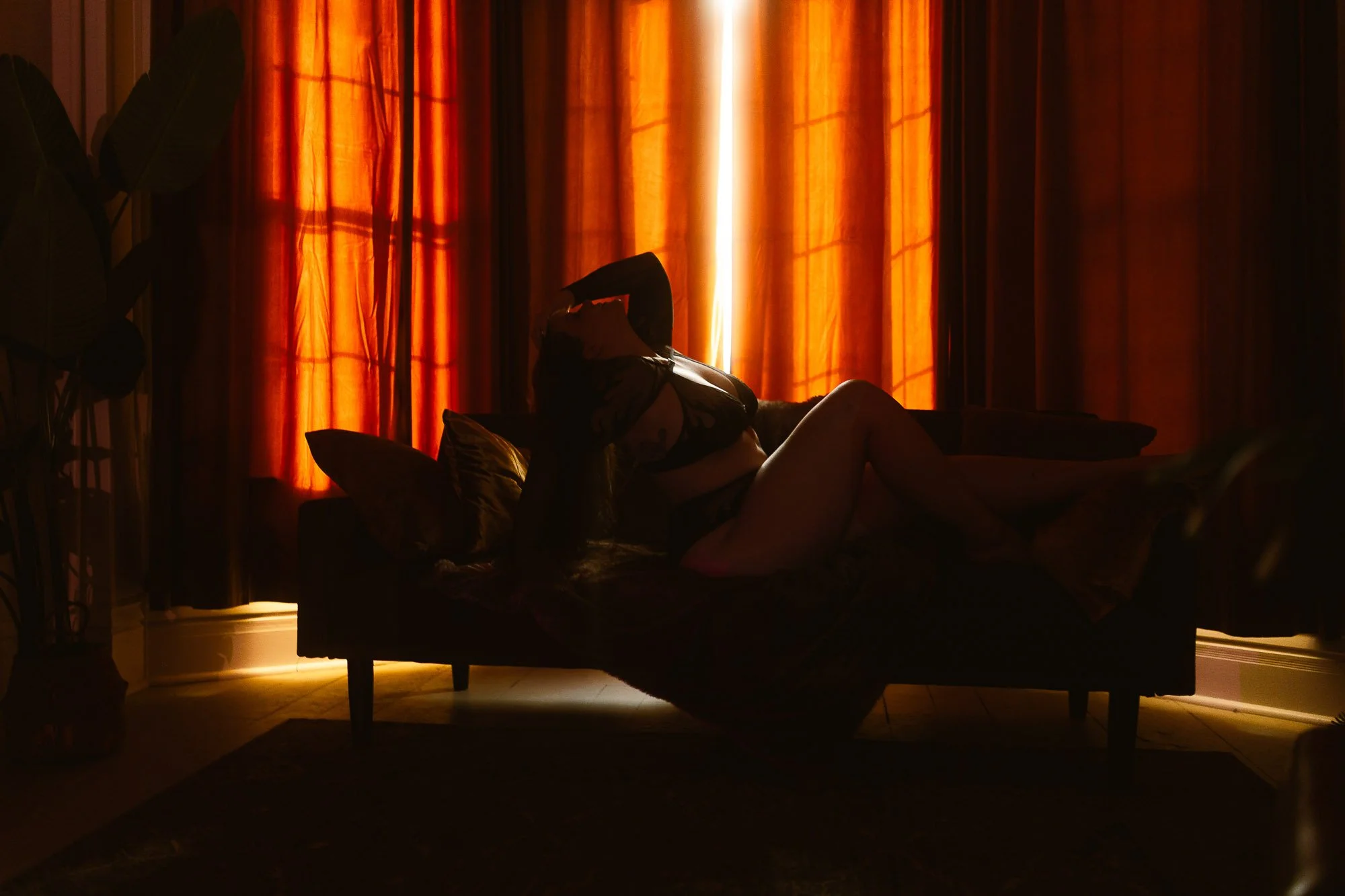 Silhouette of a woman lying on a bed in a dimly lit room with orange curtains and a potted plant to the side.