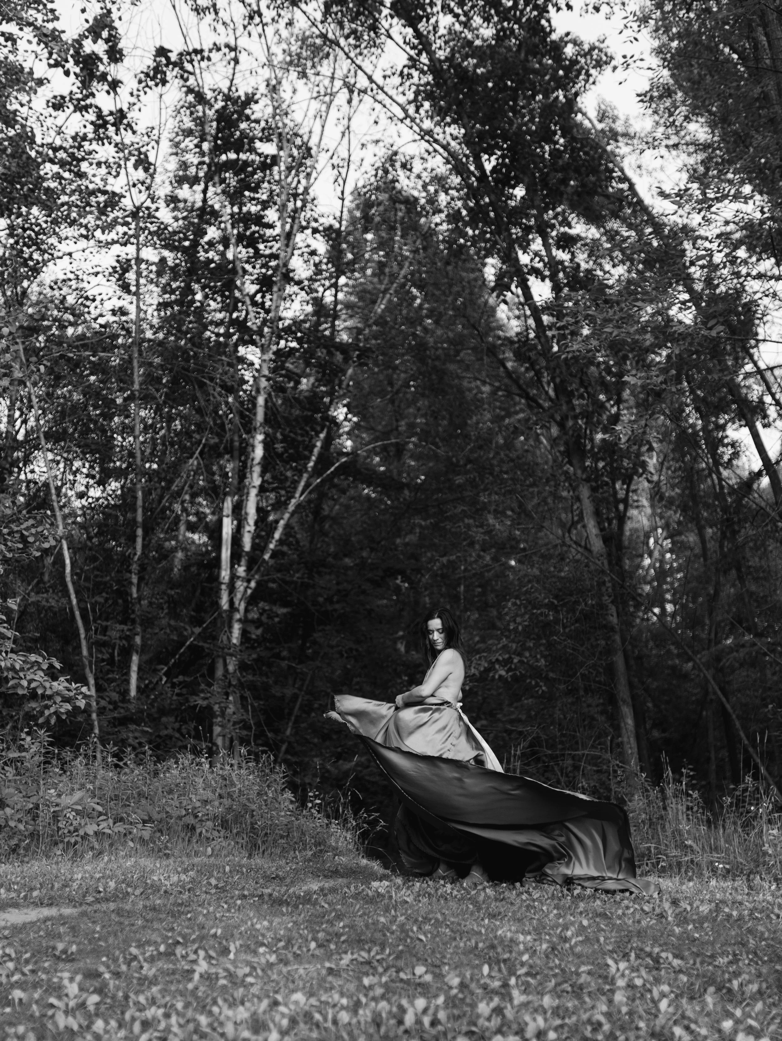 A woman sitting on a branch in a wooded area, wearing a long flowing gown, captured in black and white.