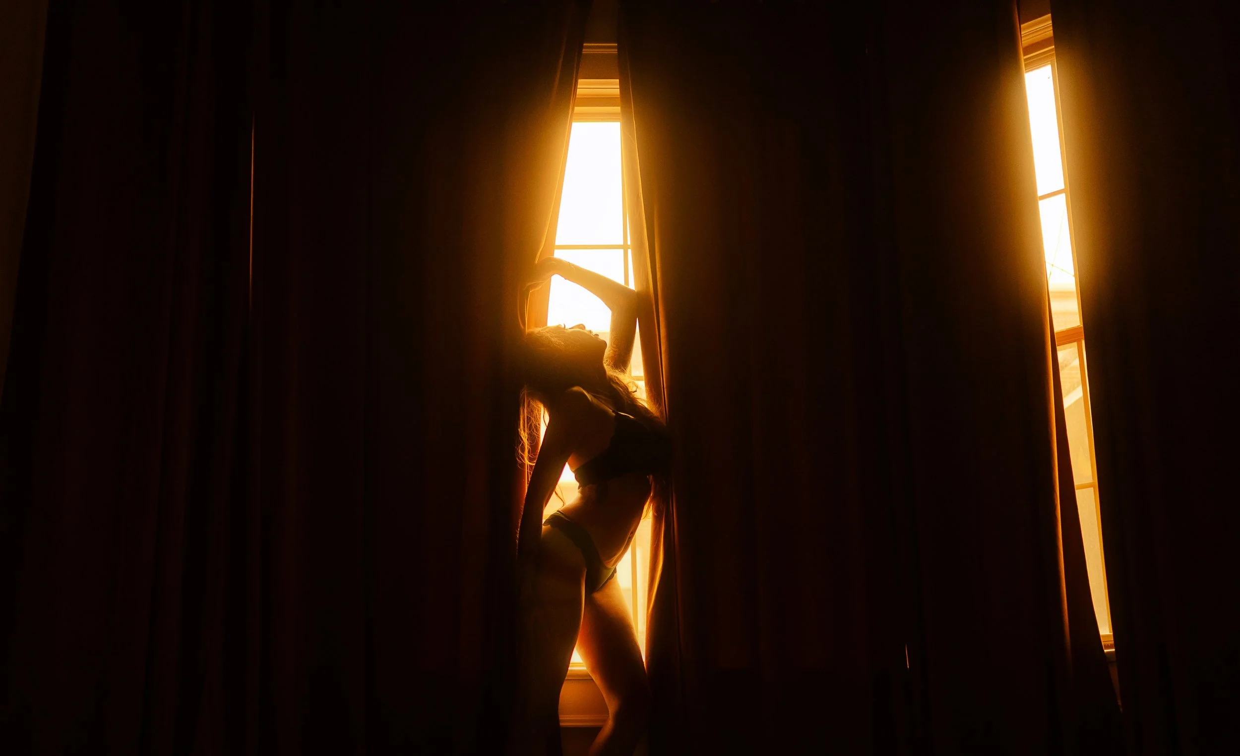 Silhouette of a woman standing by a window with sunlight streaming in, partially covered by dark curtains.