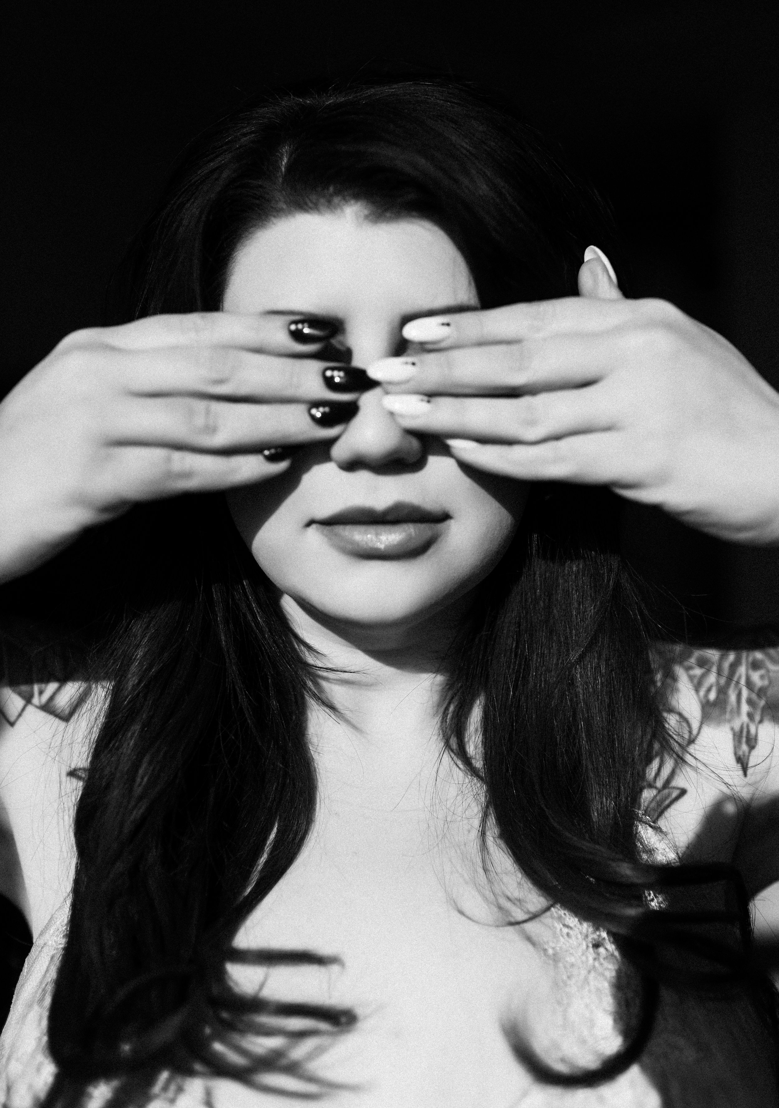 A black-and-white photo of a woman with dark hair covering her eyes with her hands, showing painted fingernails, and wearing dark lipstick.