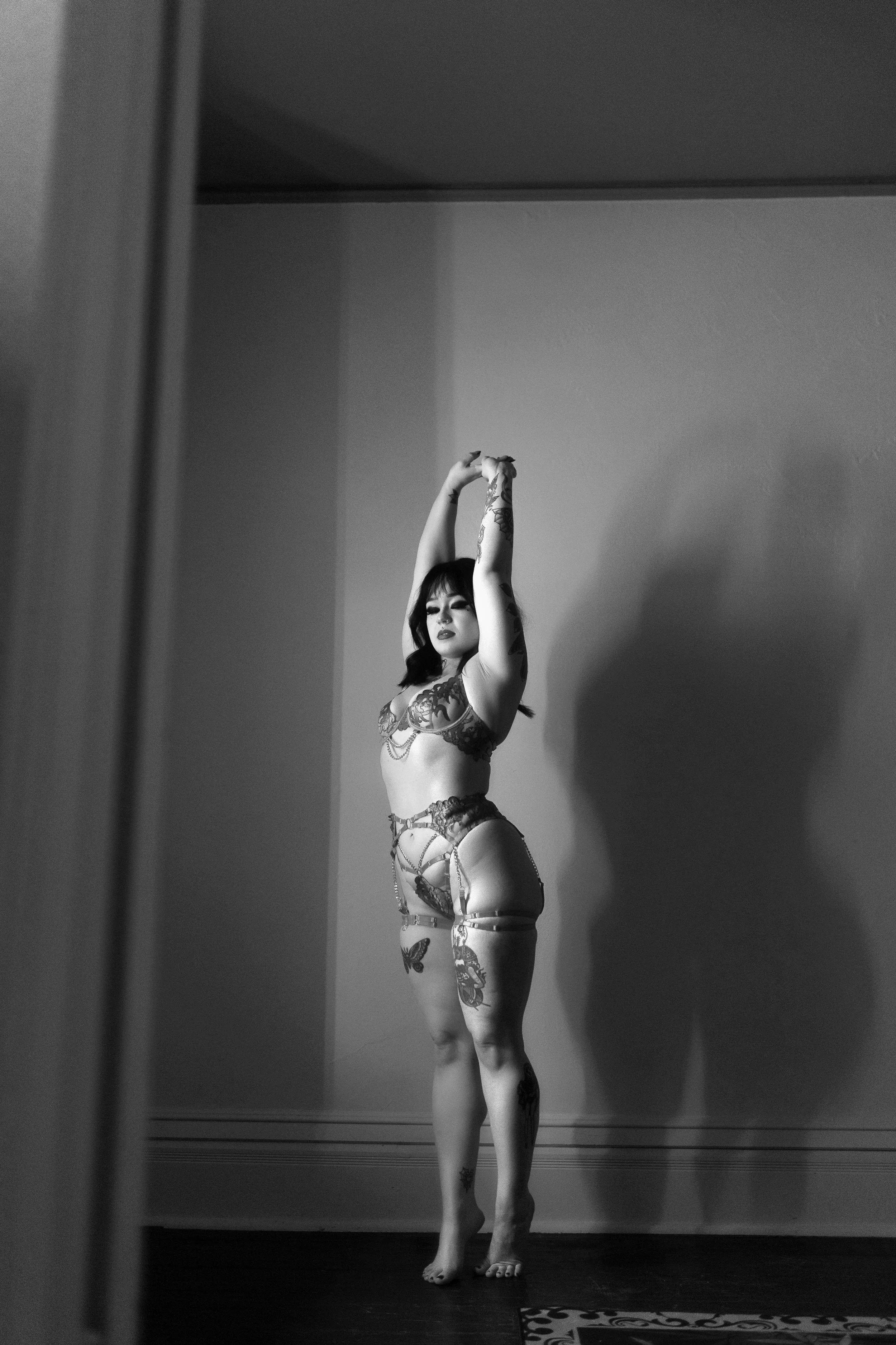 A woman standing in front of a mirror, posing with her arms raised above her head. She has tattoos on her arms and legs and is wearing lingerie. The photo is black and white.