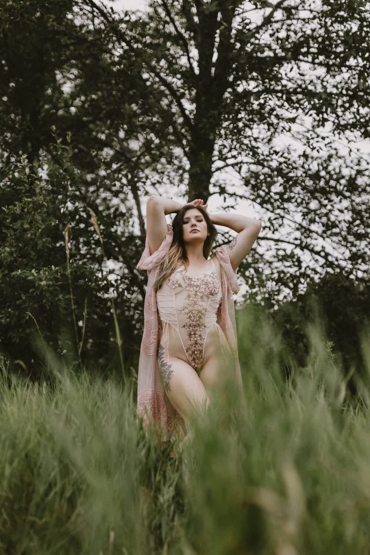 A woman with long hair standing outdoors in a grassy field, wearing a sheer, embroidered bodysuit and a light pink shawl, with her arms raised behind her head, and large trees in the background.