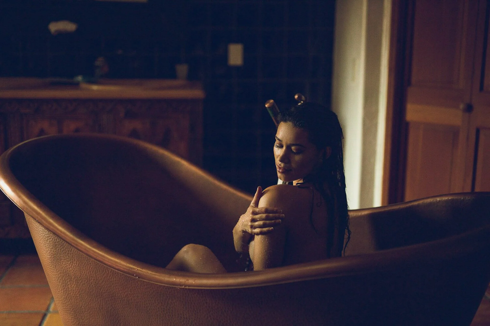 A woman in a bathtub, with a dark wood-paneled room in the background, illuminated with soft lighting.