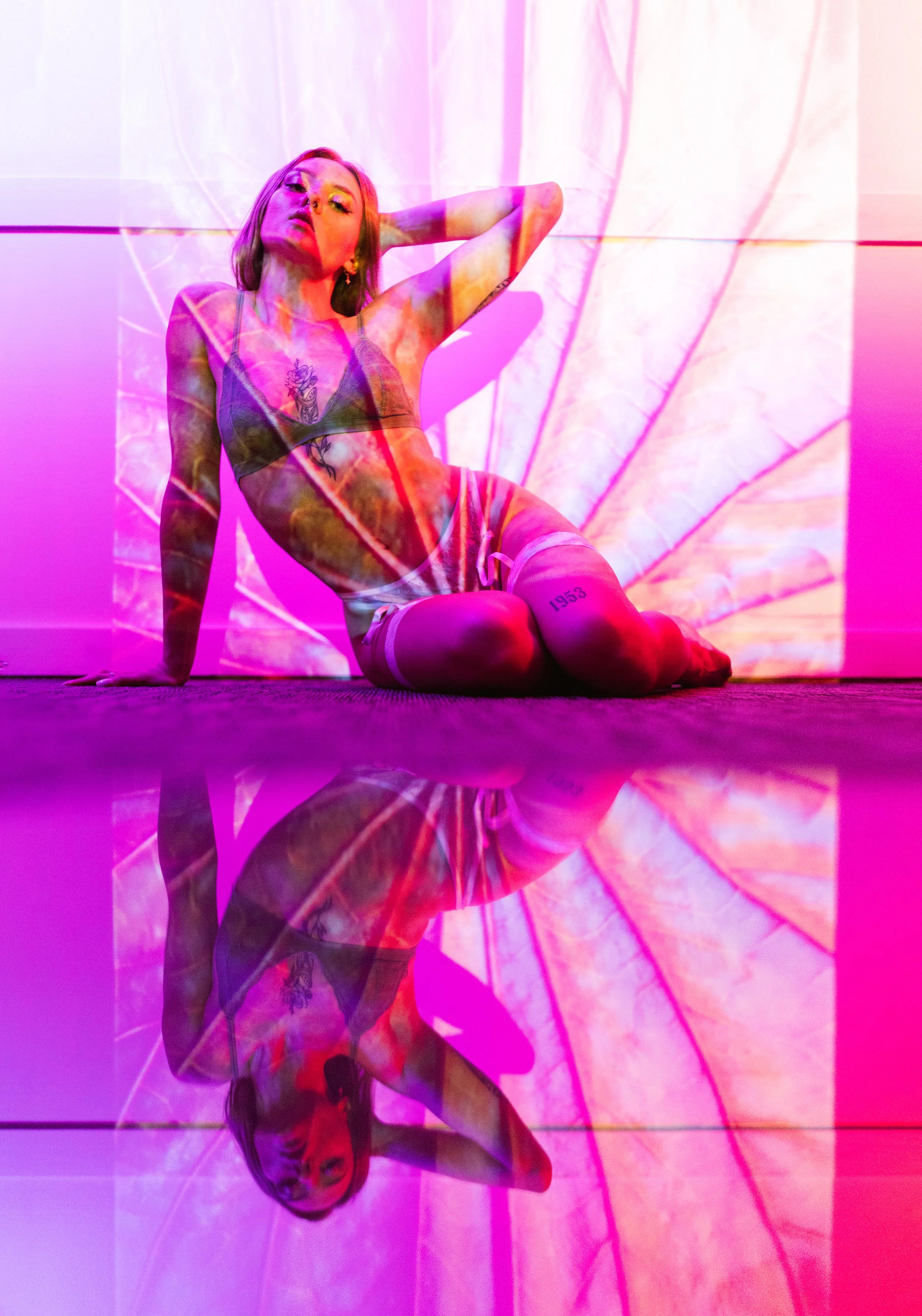 A woman with tattoos is sitting on the floor against a wall with a pink and purple digital projection. She is wearing a bra and shorts, with her left arm behind her head and her right hand on the floor. Her reflection is visible on a shiny surface be