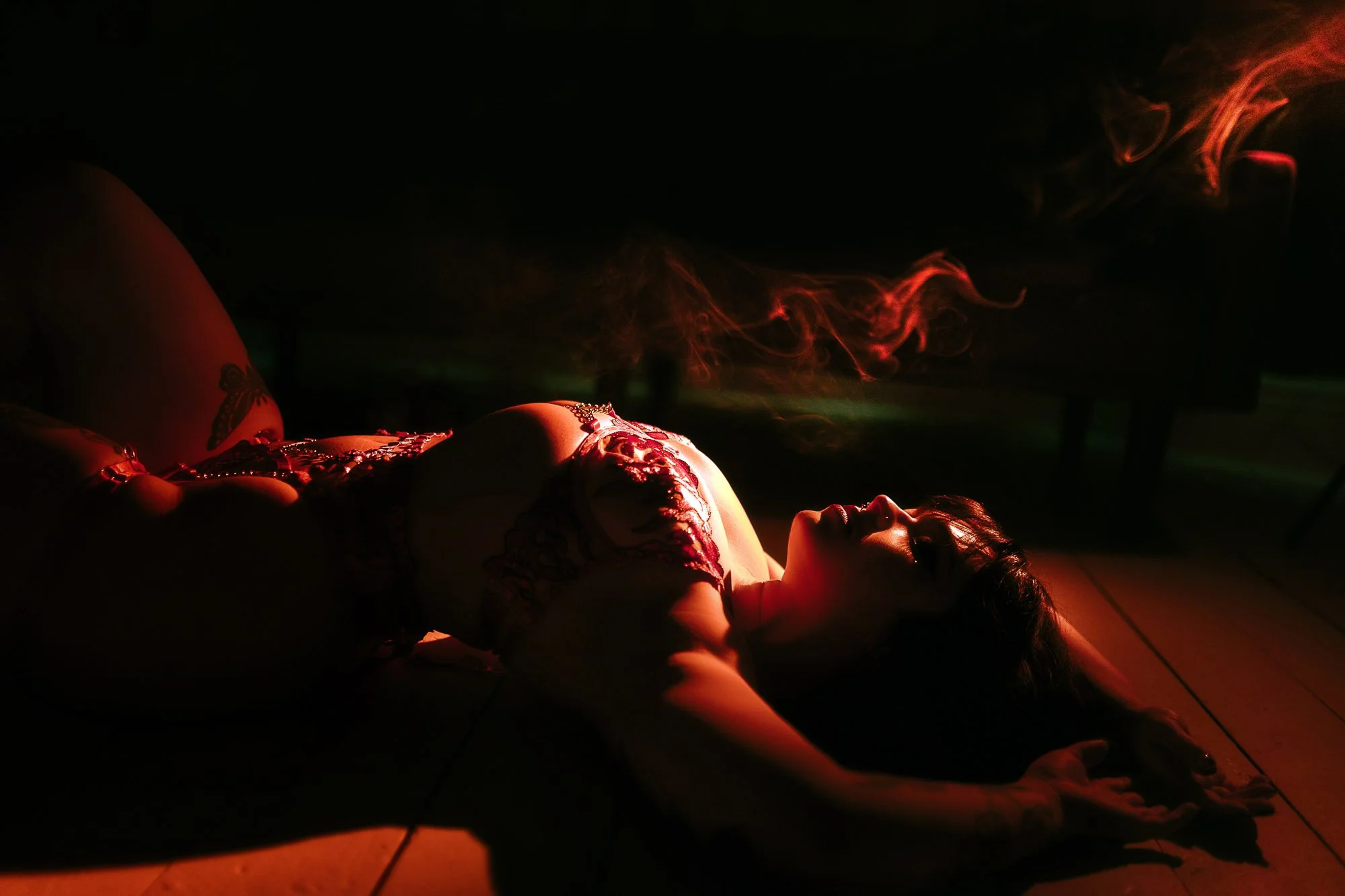 A woman lying on the floor in a dark room, illuminated by red and green lighting, with smoke in the background, wearing lingerie and jewelry.