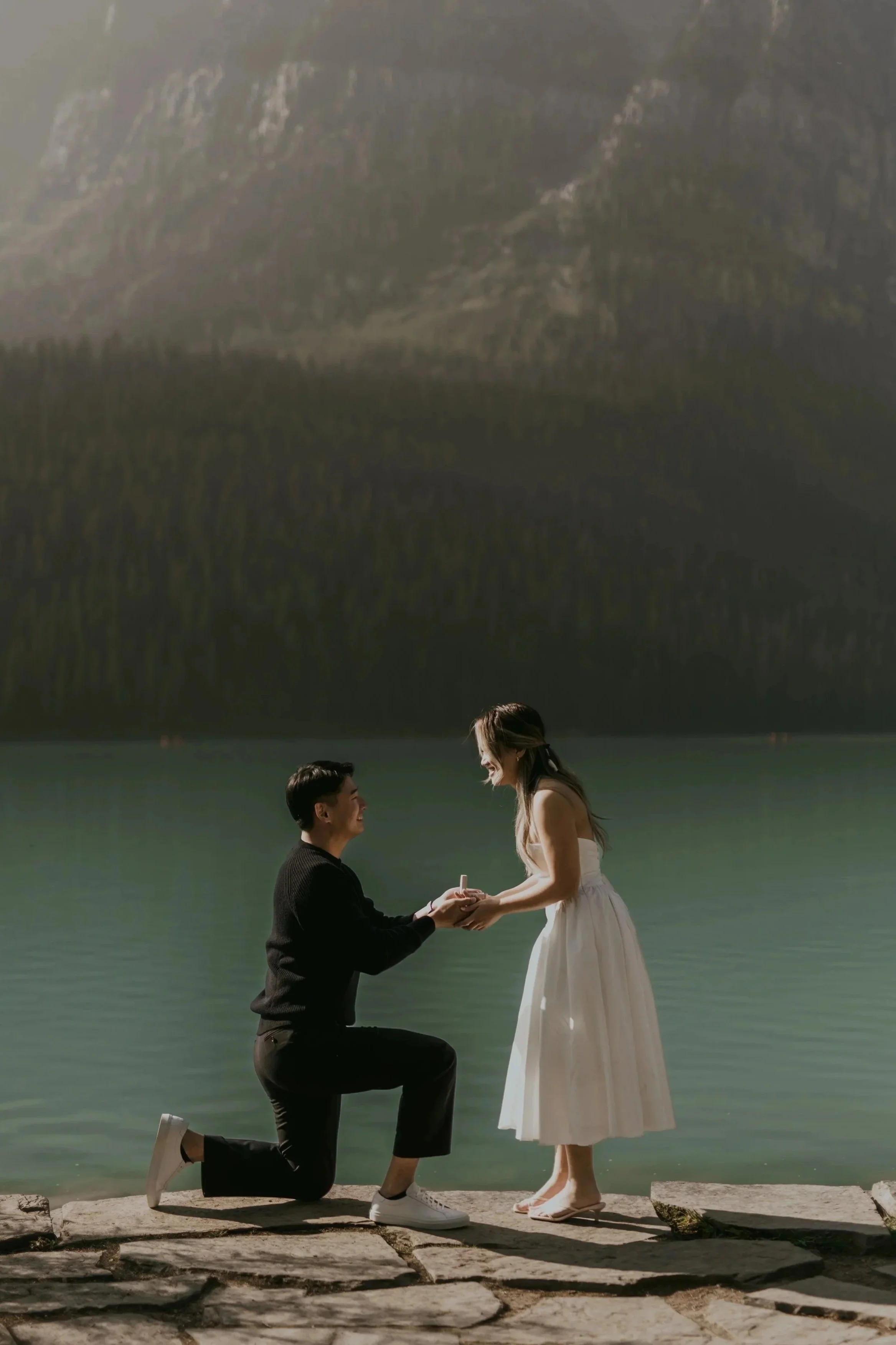 A man kneeling on one knee, holding the hands of a woman standing in front of him, by a body of water with mountains in the background. The man is wearing a black sweater and black pants, while the woman is dressed in a white dress.