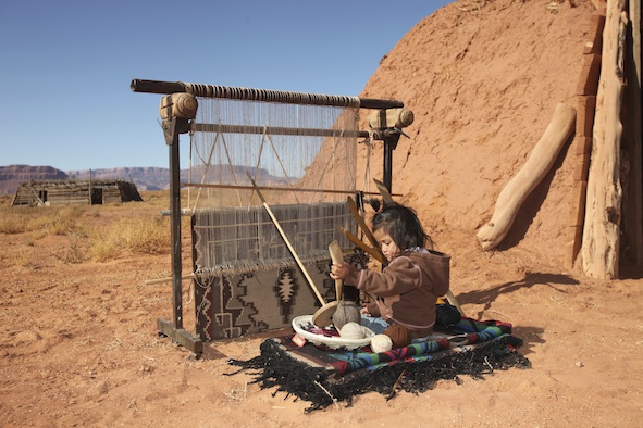 A little Navajo girl sitting at her mother's loom. This loom is outside a traditional Navajo home called a hogan, and there is another one in the background. However, most Navajo live in modern houses. ©Getty Images