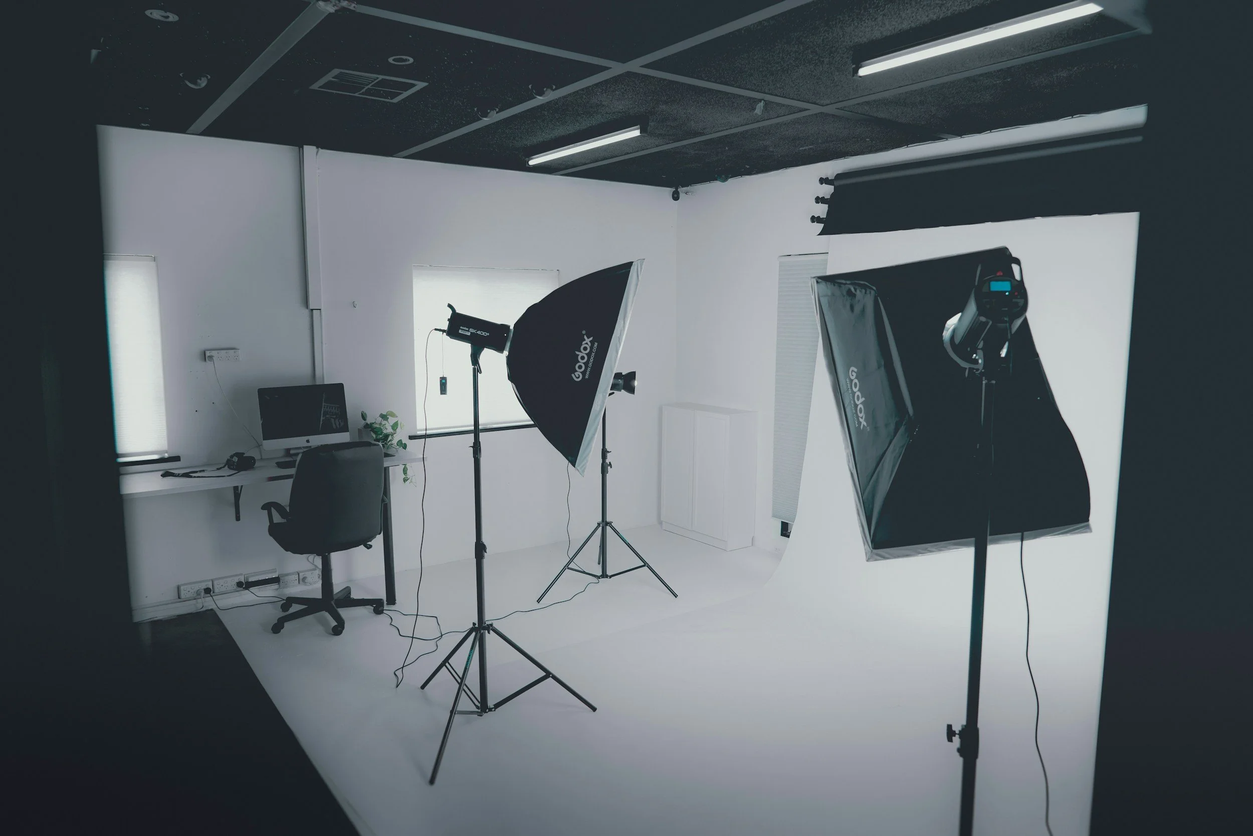 Photography studio with two large softbox lights, a small white desk with a computer, a black office chair, and a white backdrop in a professional setup.