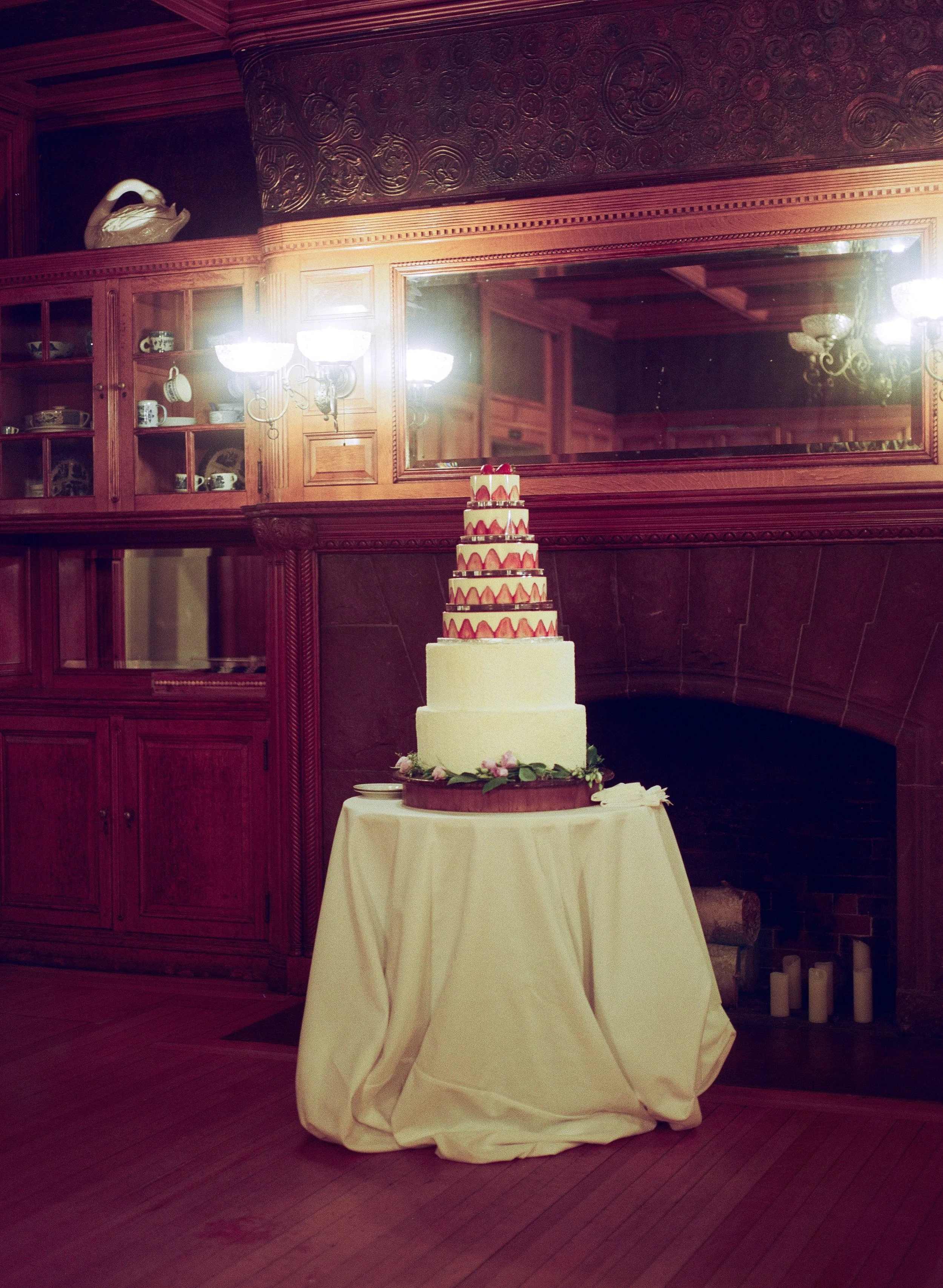 A multi-tiered wedding cake on a table with a cream-colored tablecloth, set in a room with wooden paneling, a large mirror, and a decorative ceramic swan on a shelf.