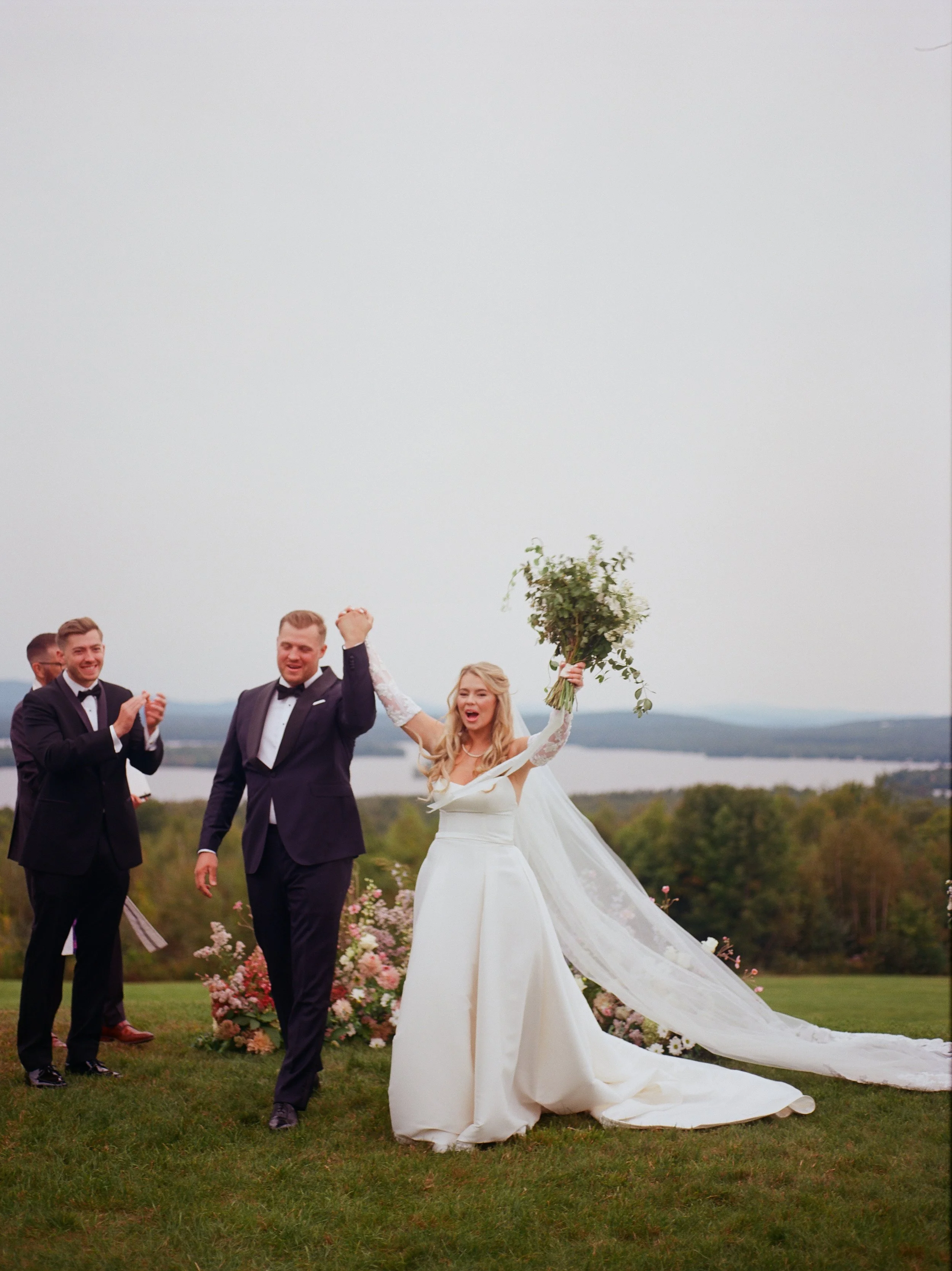 A bride and groom celebrating their wedding outdoors with friends, holding a bouquet of flowers, with a scenic landscape and a cloudy sky in the background.