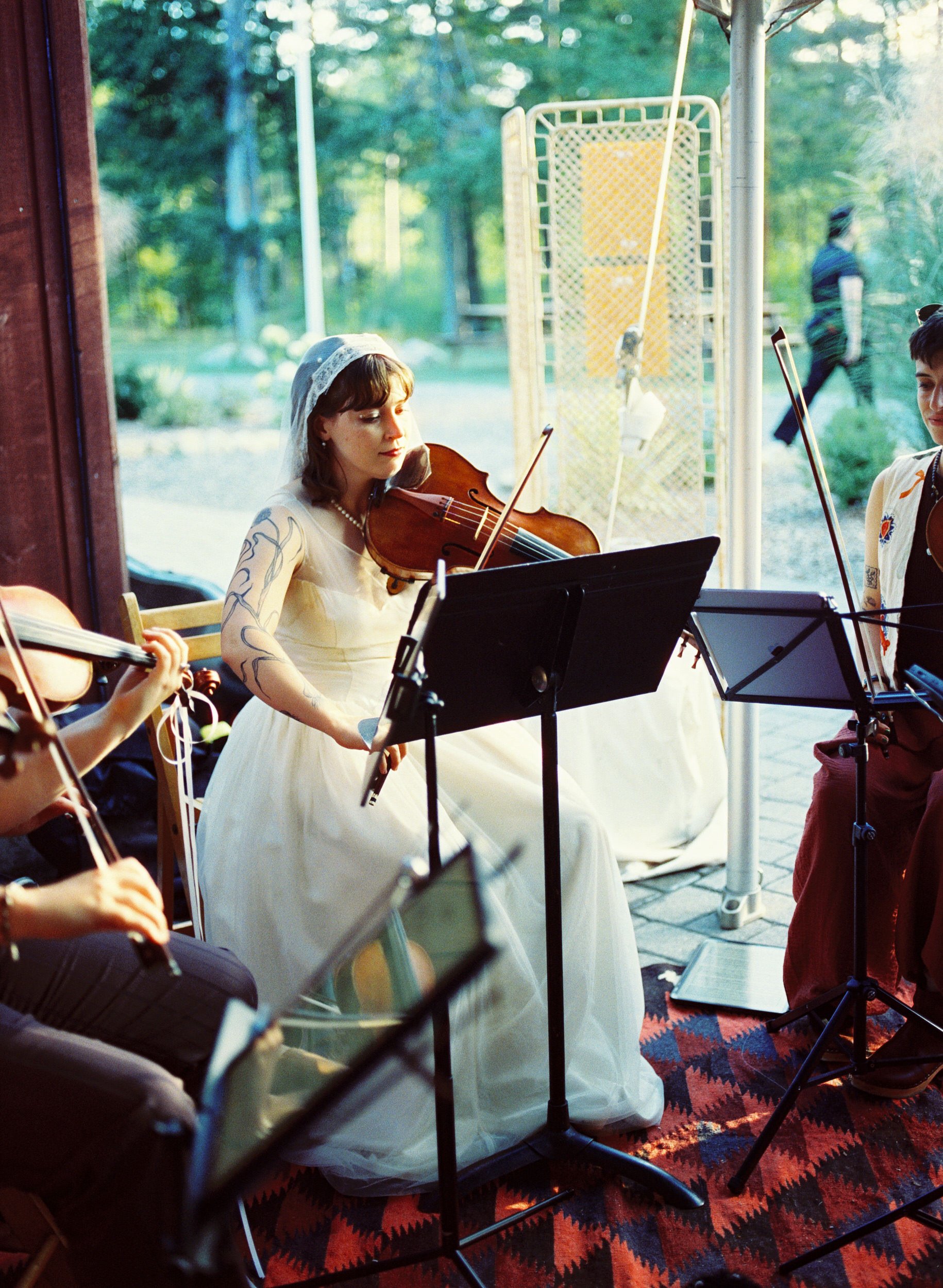 A woman in a white dress and veil playing a violin, sitting on a chair, surrounded by other musicians who are also playing string instruments. The scene appears to be an outdoor performance with greenery visible in the background.