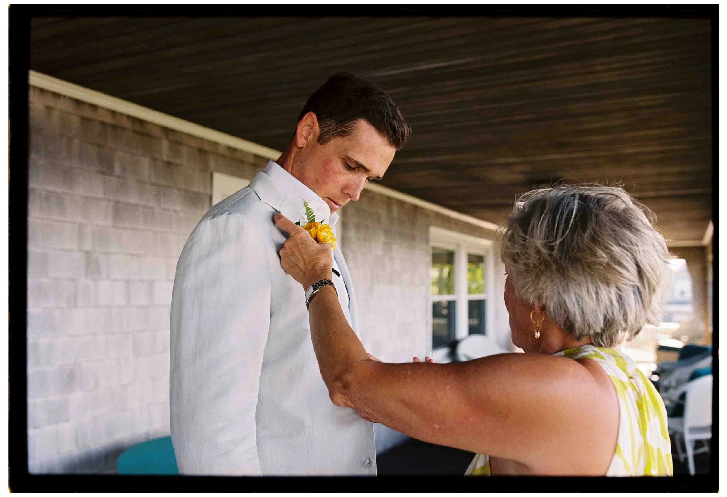 mother in yellow floral dress pins boutonniere on son's wedding suit jacket