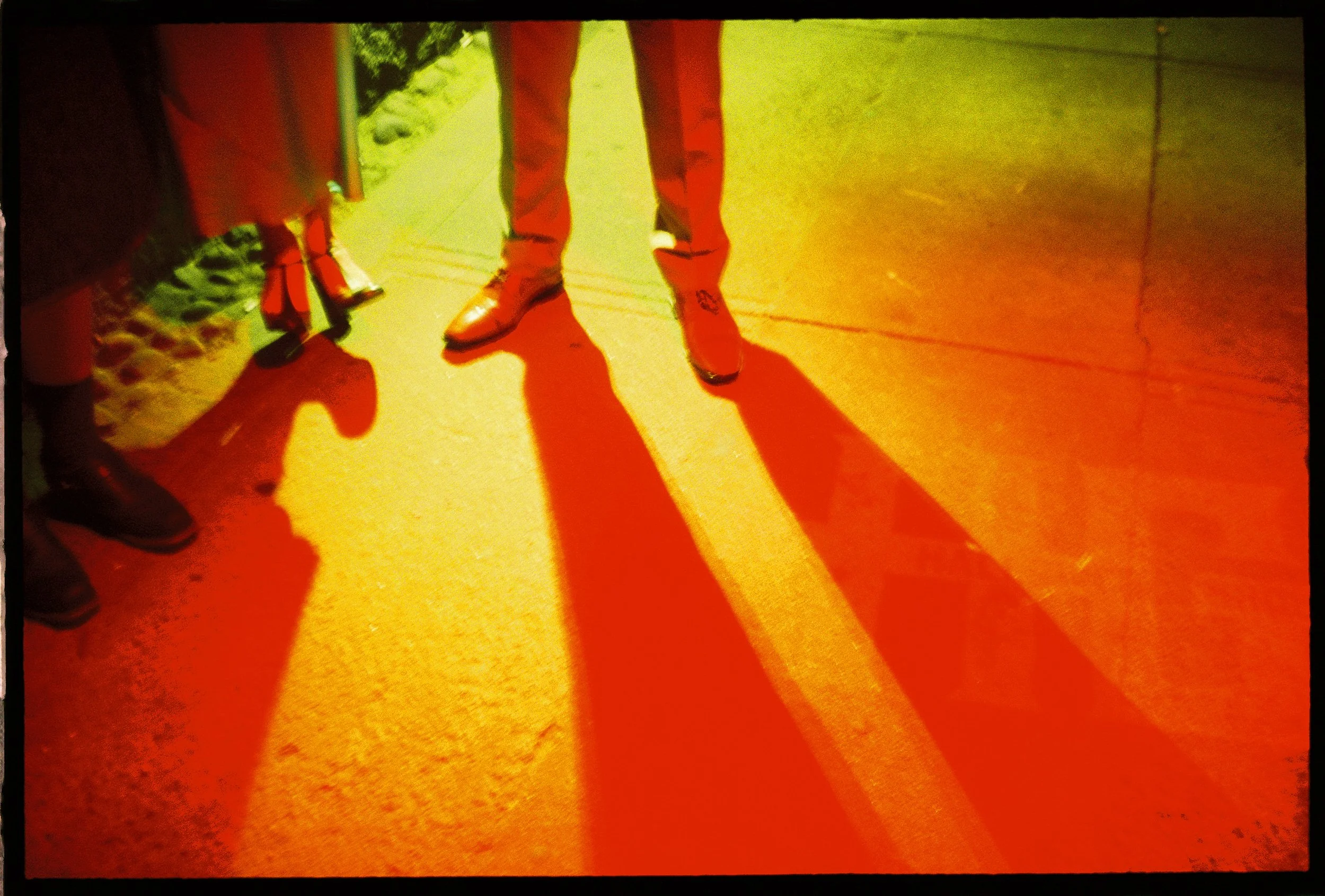 red and orange saturated shadows of peoples feet outside at night with one set of shoes balancing on the heels outside of a wedding after party