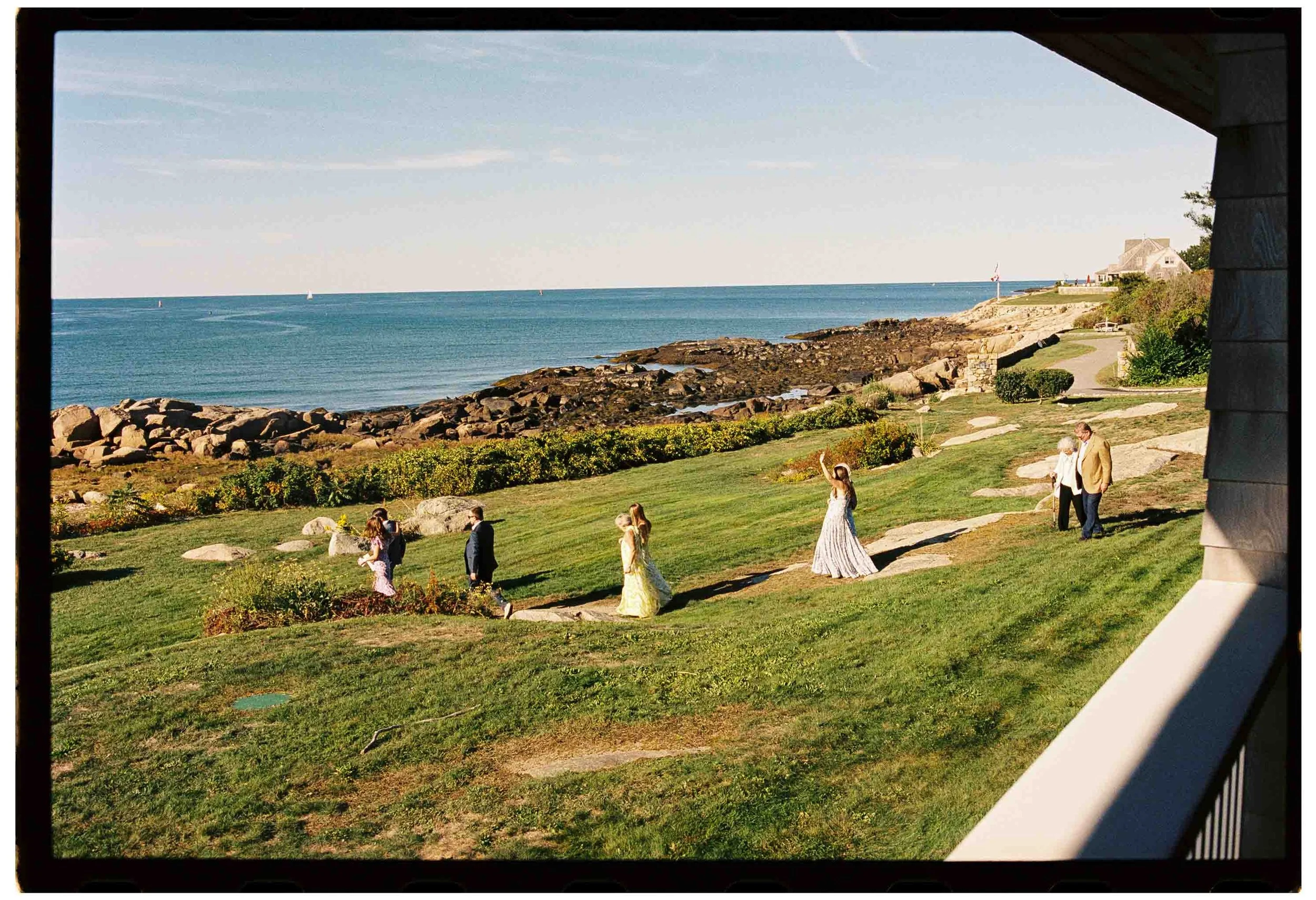 A scenic view through a window overlooking a grassy yard with people, some dressed in formal attire, walking and a woman waving. In the background, there is a rocky shoreline and the ocean with sailboats, and a house with a flag on a pole.