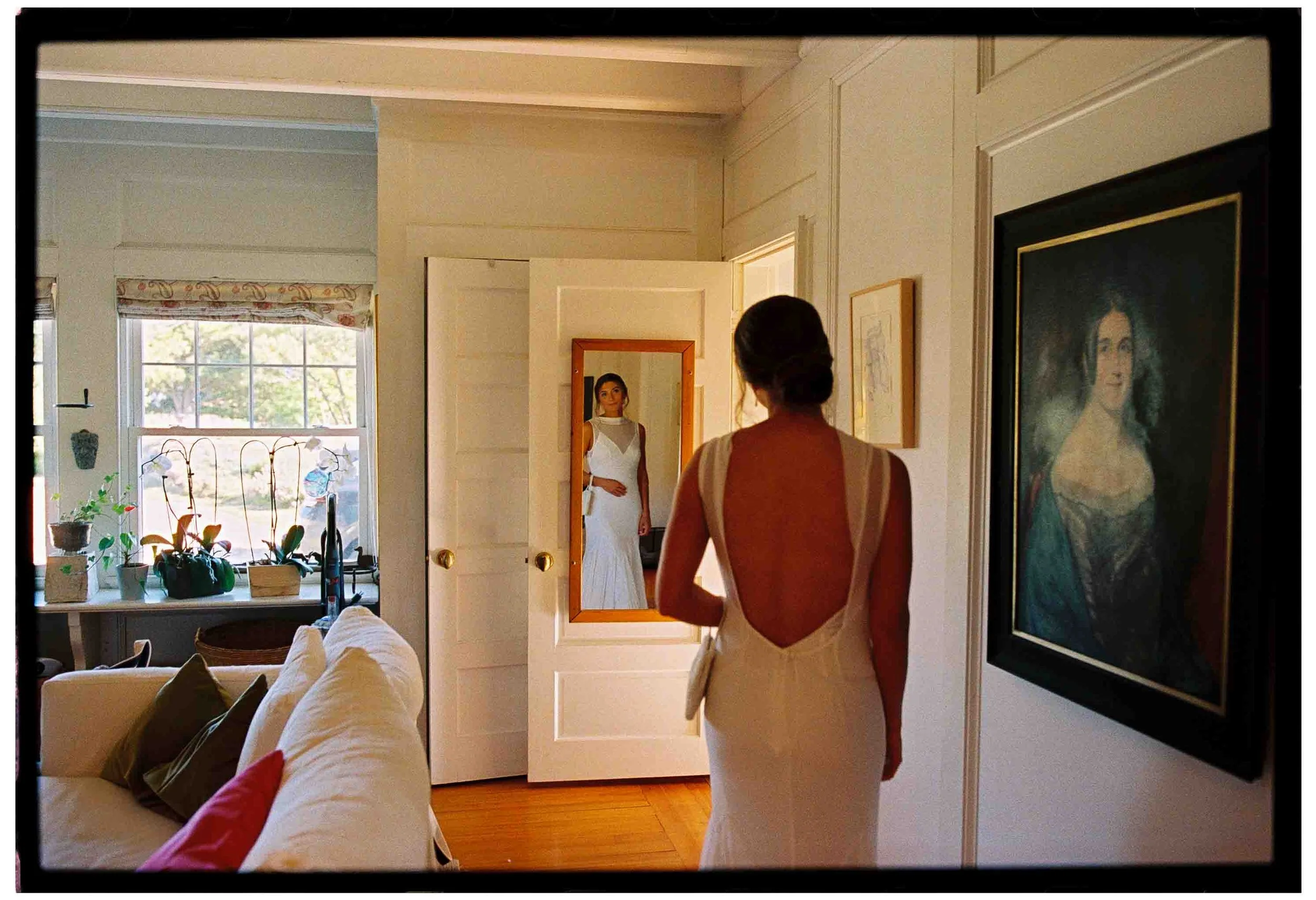 bride admires her outfit in mirrior in ancestral home with family member's painting in the frame