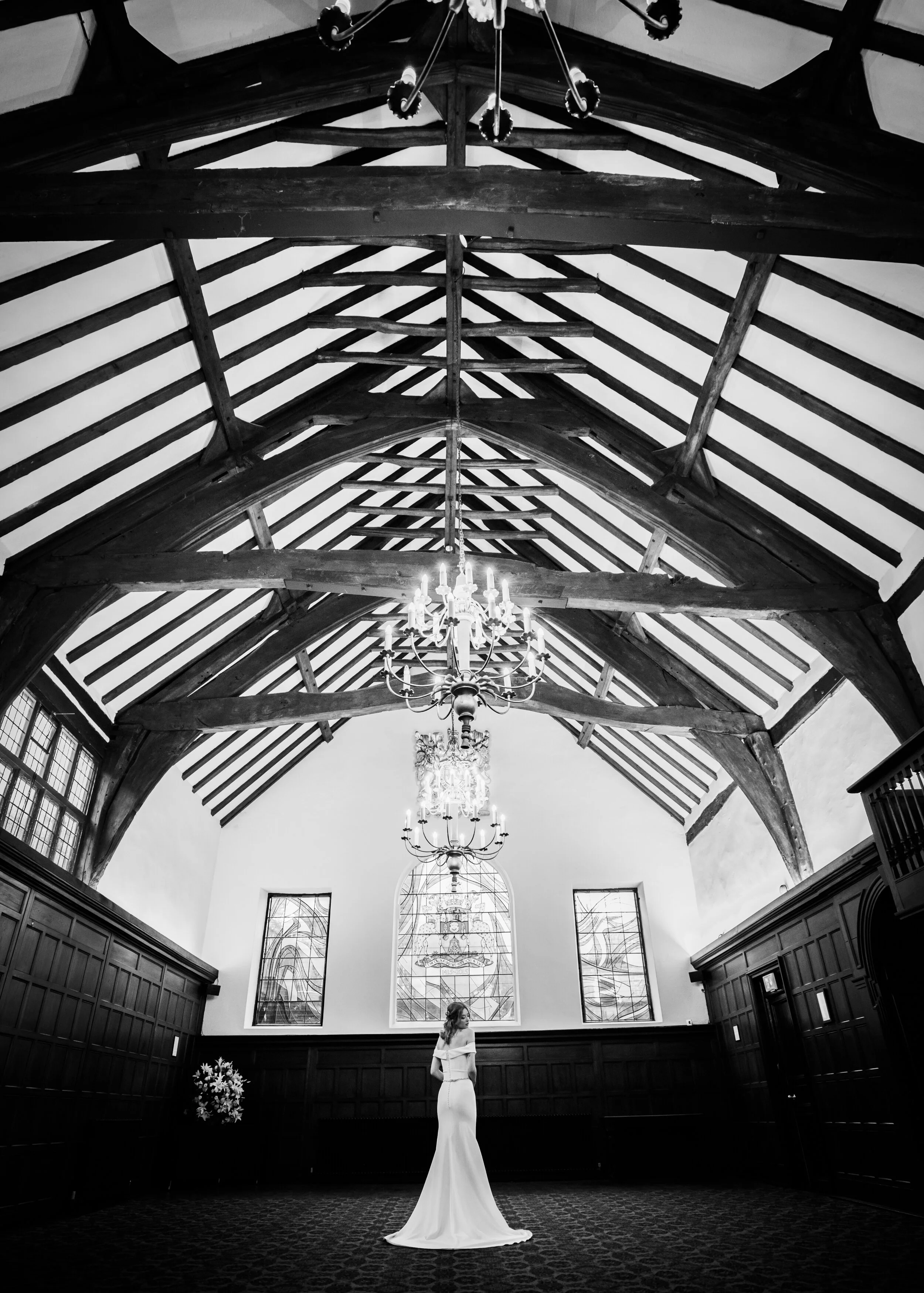 A magnificent medieval timbered hall with stained glass windows and chandeliers, empty except for a bride in a stunning off the shoulder gown