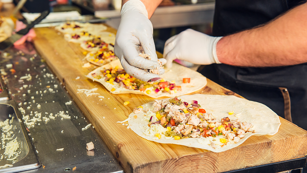 A food truck cook prepping several tacos with sanity gloves on.