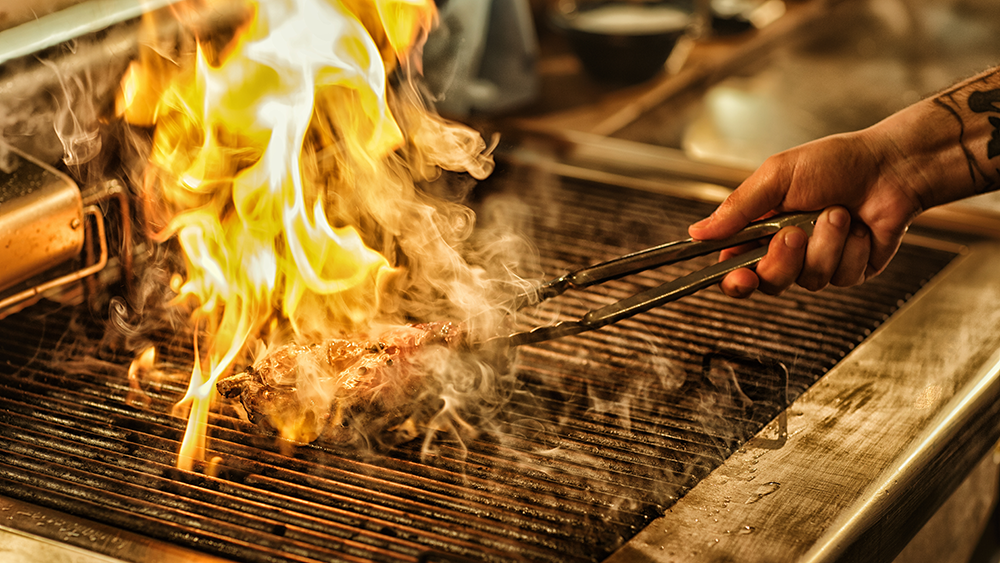 Close up of a chef cooking meat over an open fire. A flame sizzles over it.