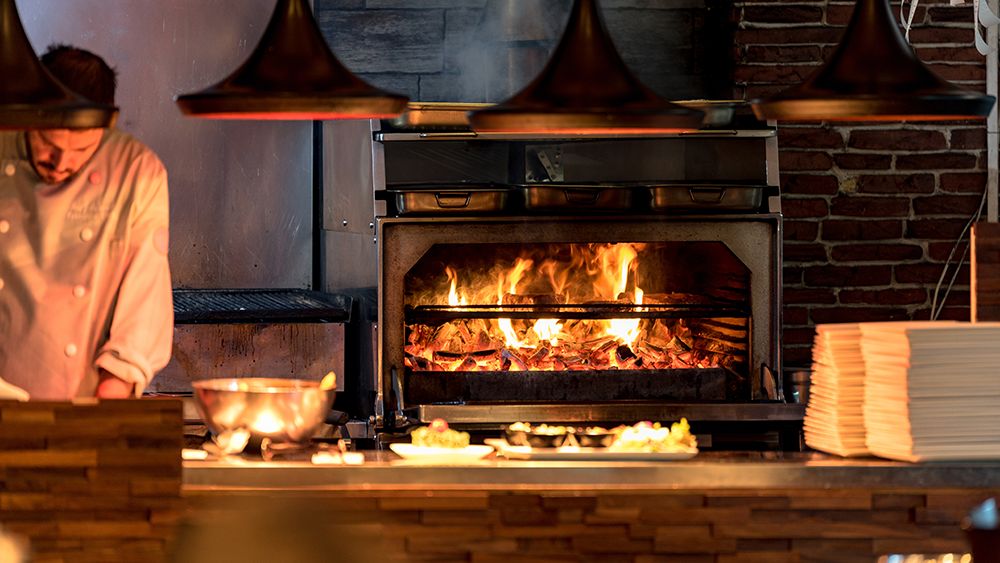 A restaurant chef works while in an orange glow from a wood fire stove nearby