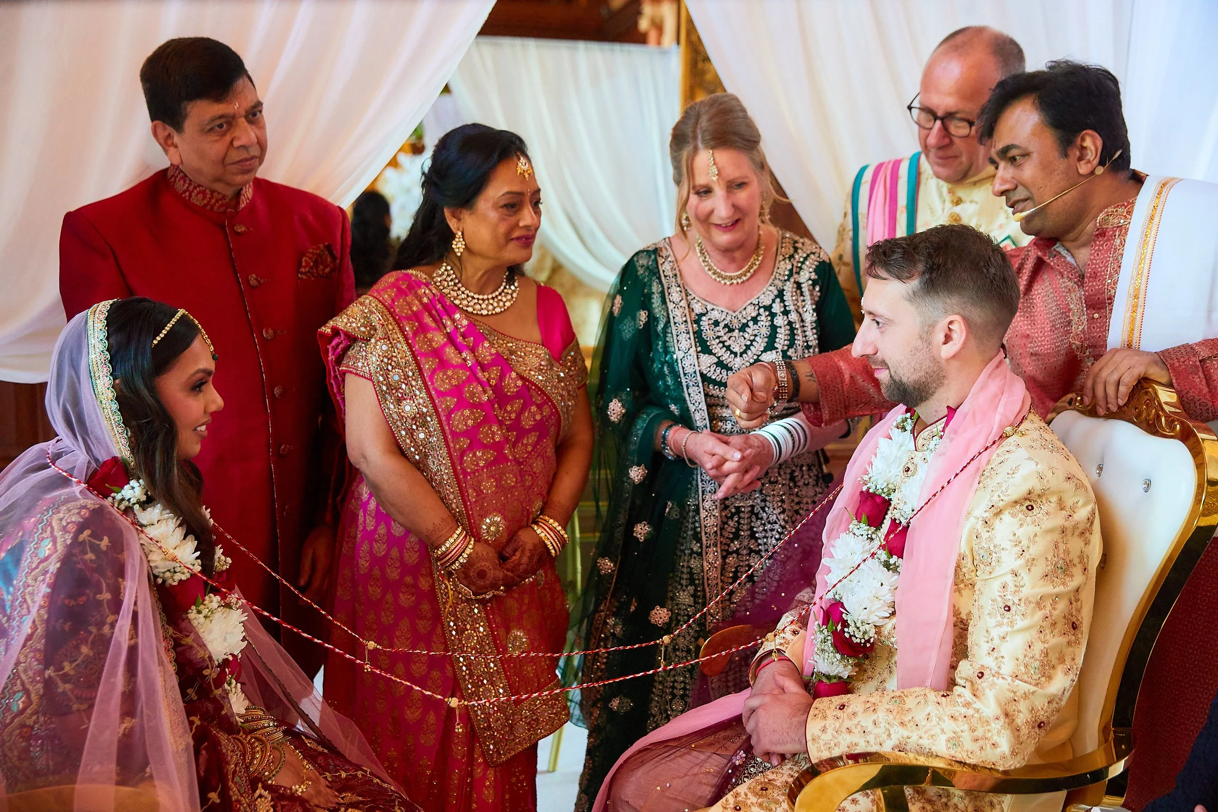 People dressed in traditional Indian wedding attire gather around a seated groom, who is sitting on a throne-like chair, during a wedding ceremony.