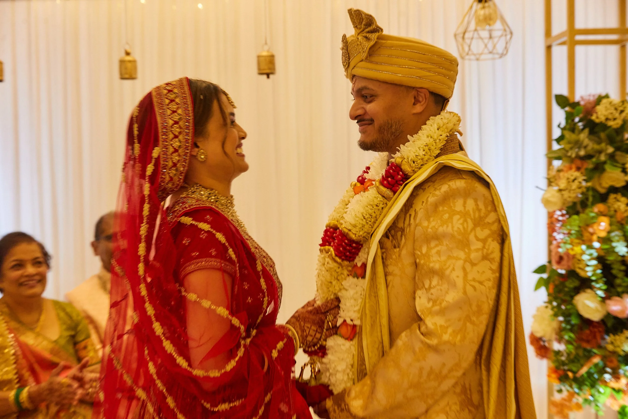 A bride and groom in wedding attire smiling and standing close during a wedding ceremony, with women in traditional dresses in the background.