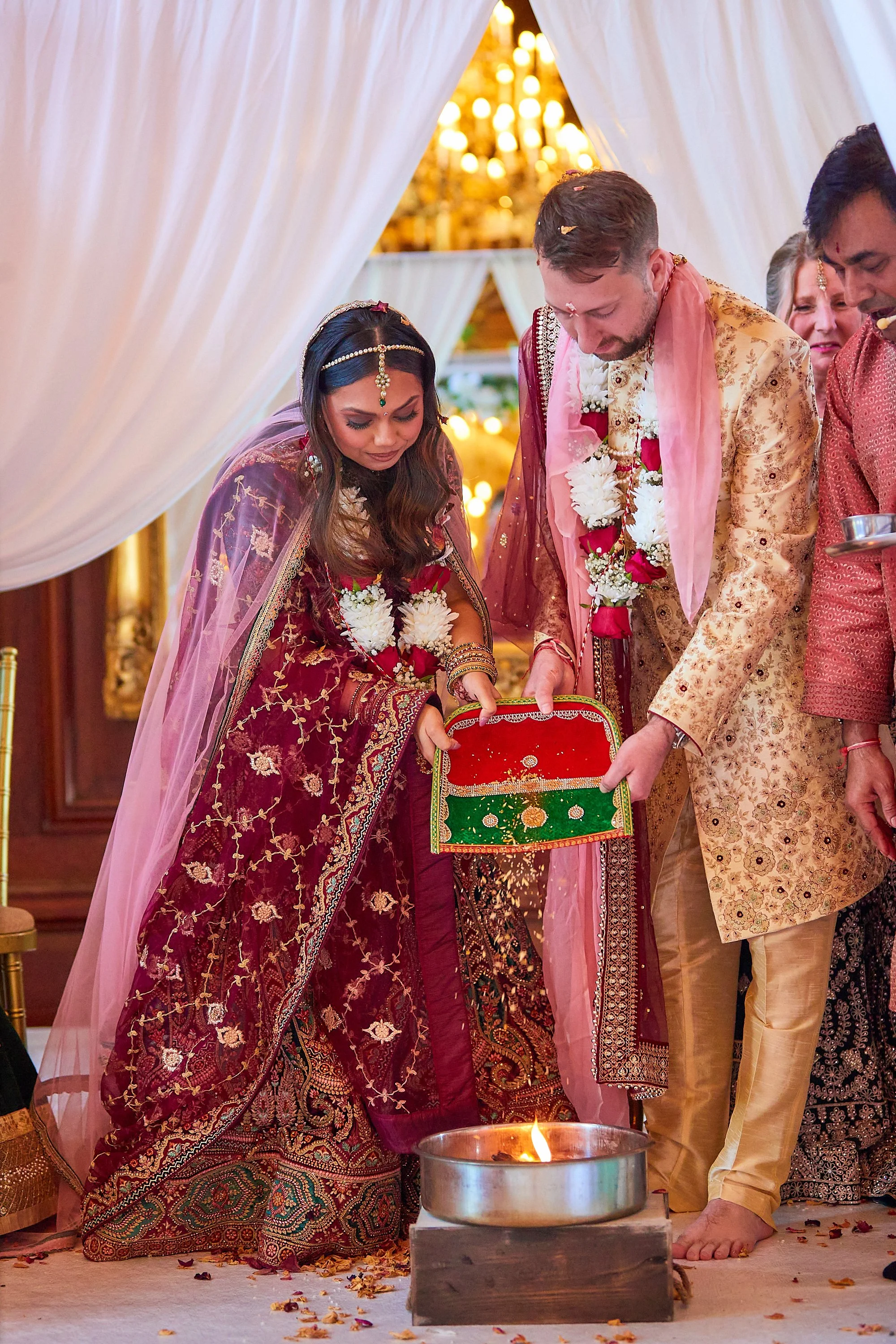 Indian bride and groom participate in a traditional wedding ceremony, with the bride holding a decorated box over a fire altar, surrounded by family and friends in wedding attire.