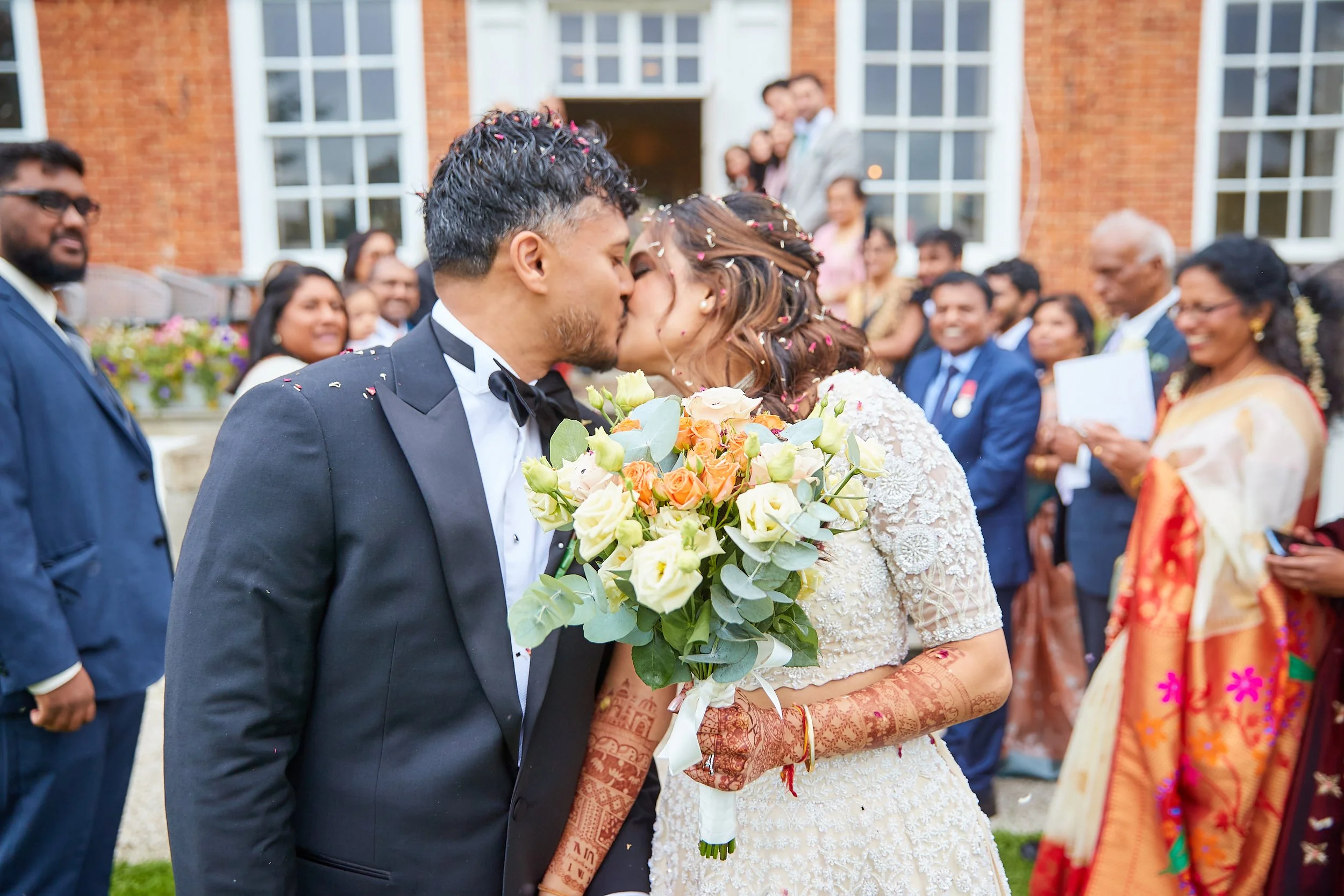 A newlywed couple sharing a kiss at their wedding ceremony, with the bride holding a bouquet of flowers and guests smiling in the background outside a brick building.