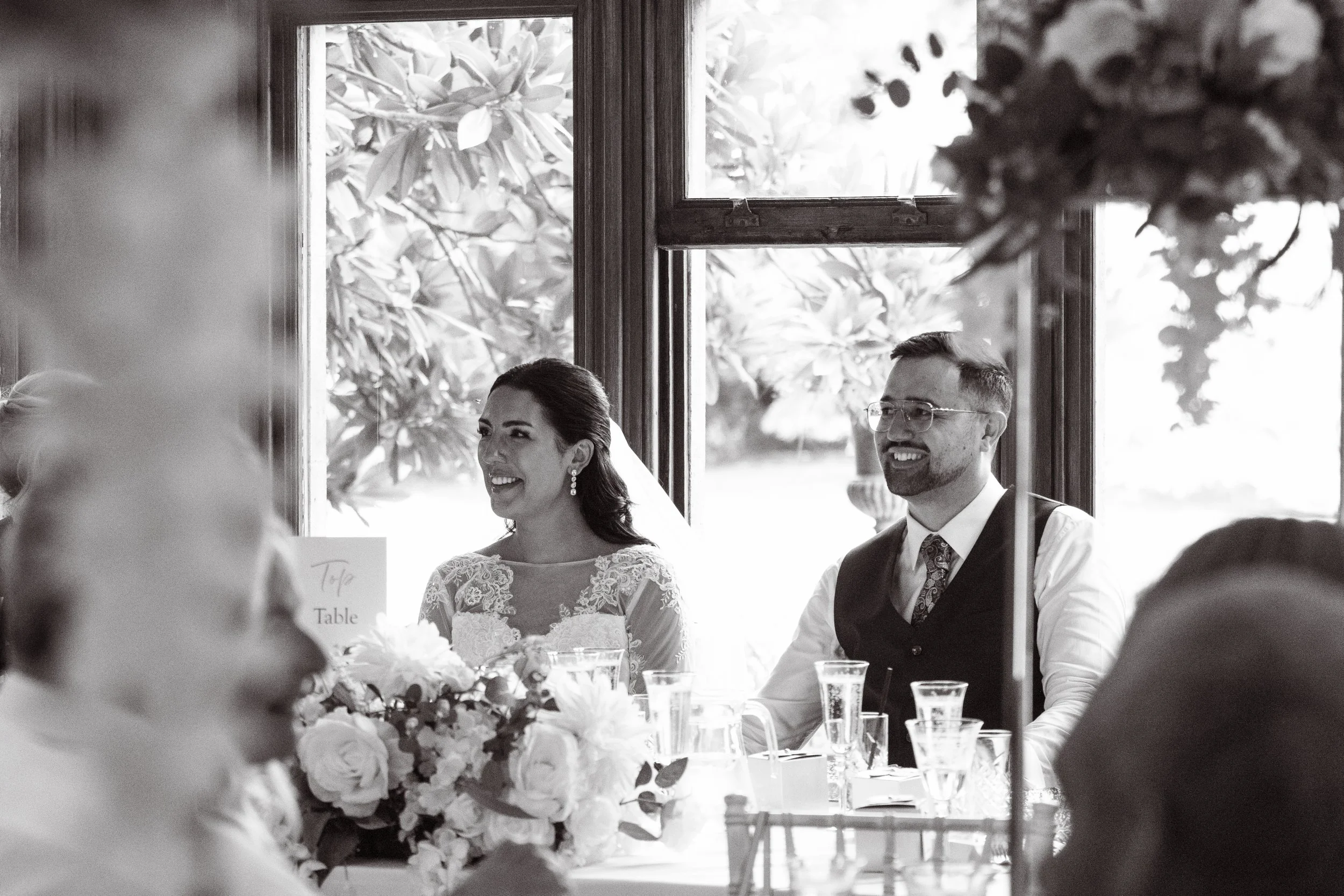 Black and white photo of a bride and groom sitting at a wedding reception table, smiling and laughing. The bride is wearing a lace wedding dress, and the groom is in a vest and tie. They are near a window with trees outside, and the table has bouquet