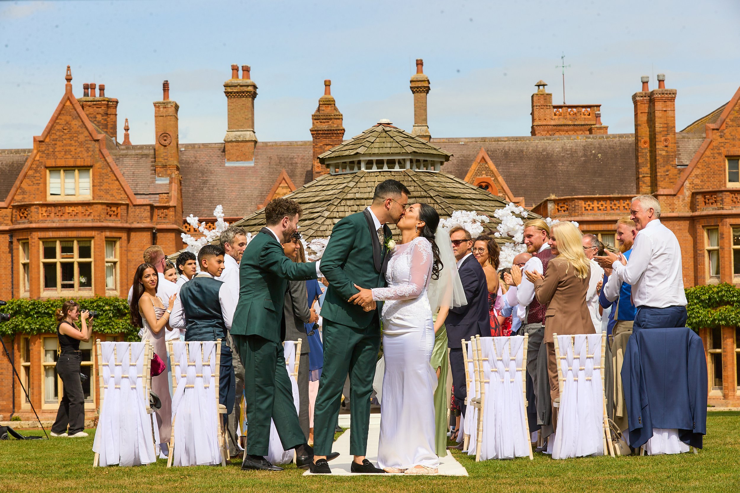 A wedding ceremony outdoors with a bride and groom kissing, surrounded by guests, on a lush green lawn in front of a large historic building with brick architecture.