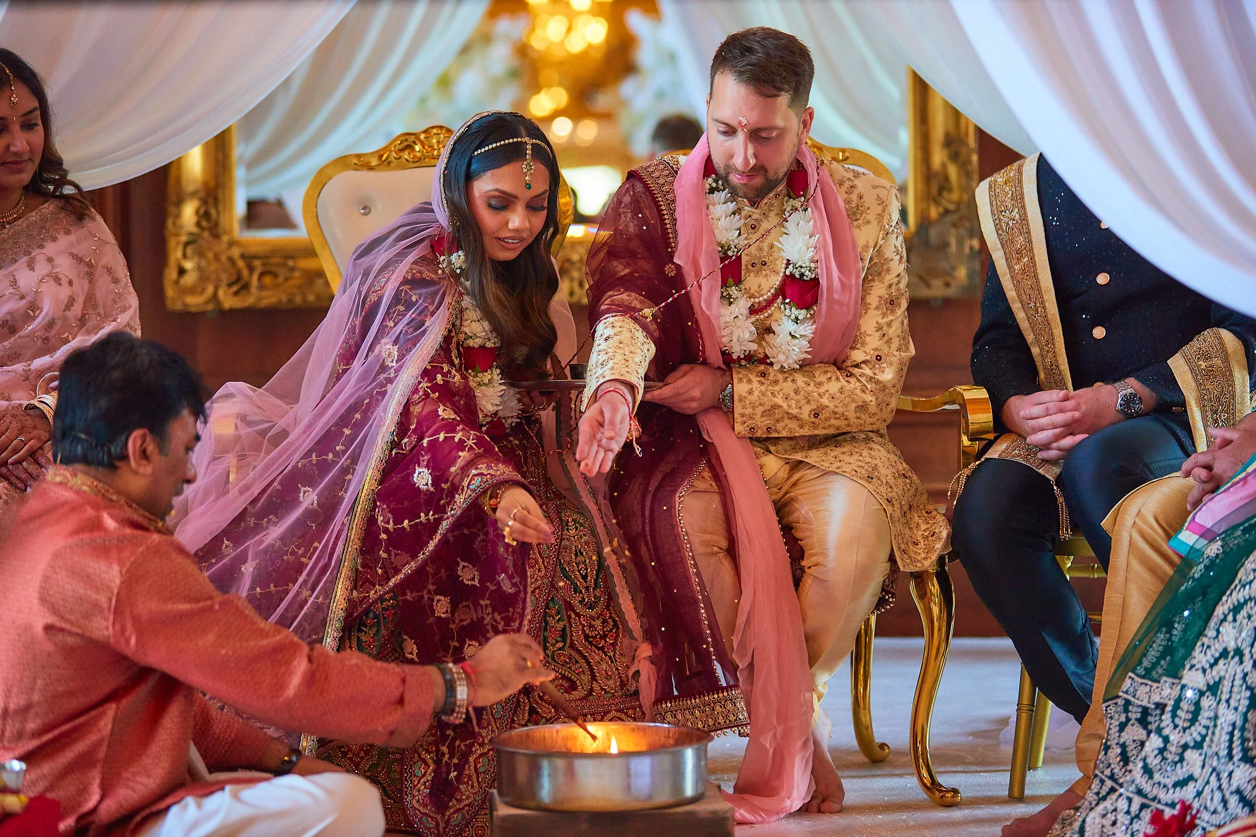 Indian wedding ceremony with bride and groom dressed in traditional attire, lighting a ritual fire, surrounded by family members.