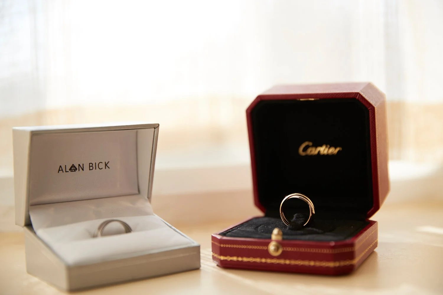 Two jewelry boxes, one from Alain Bick and the other from Cartier, each holding rings, placed on a light-colored wooden surface near a window.