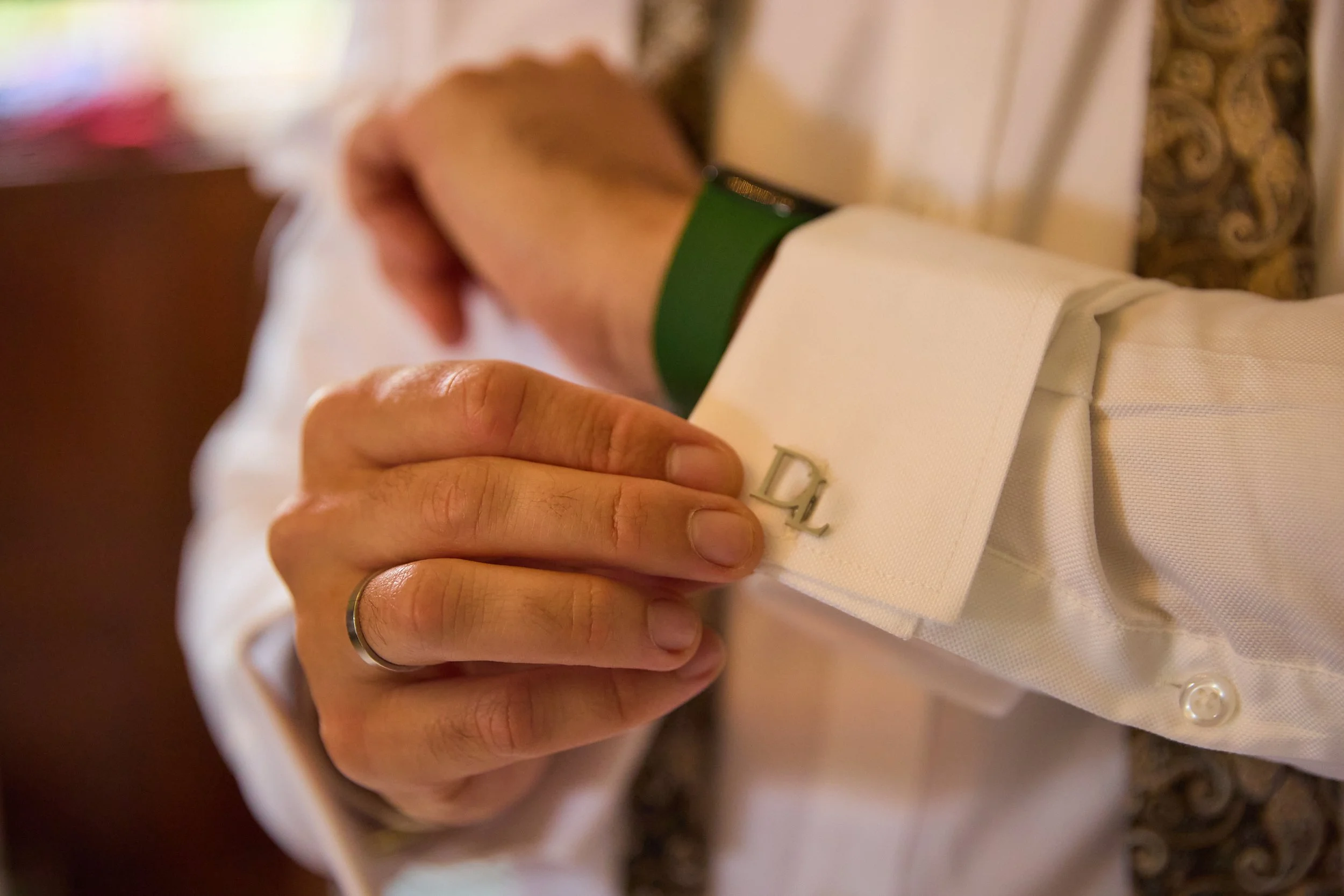 Close-up of a man adjusting his cufflinks on a white dress shirt. The cufflinks are silver and engraved with the letter 'D'. The man wears a gold wedding band on his ring finger and a green wristband.