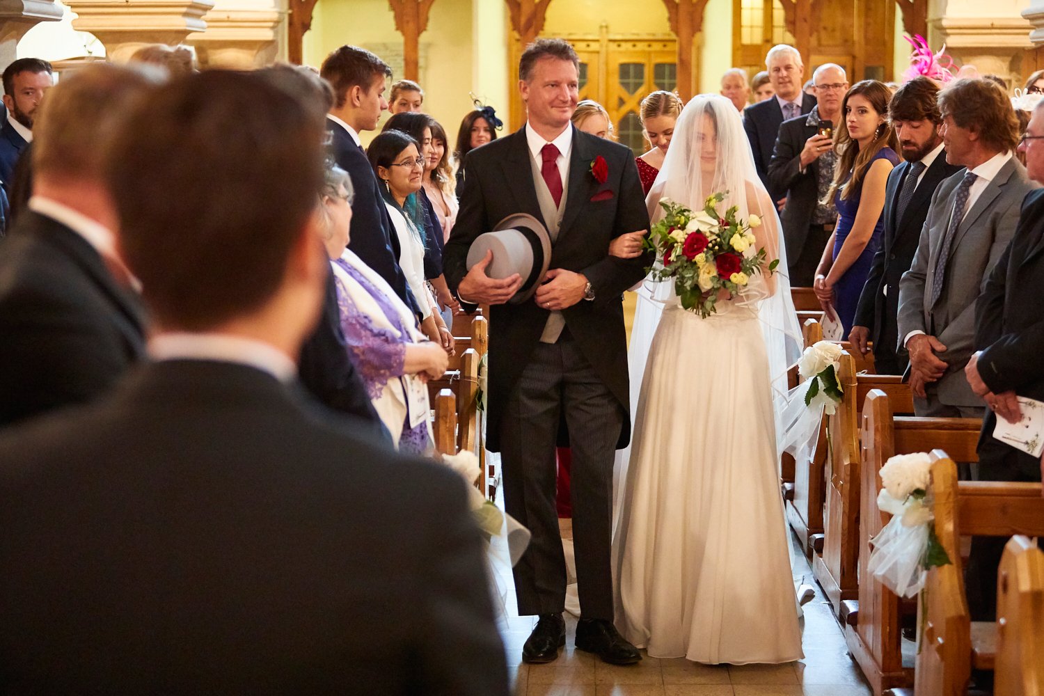 Bride walking down the aisle with her father at her wedding, holding a bouquet of red, white, and yellow flowers, in a church decorated with white flowers and ribbons.