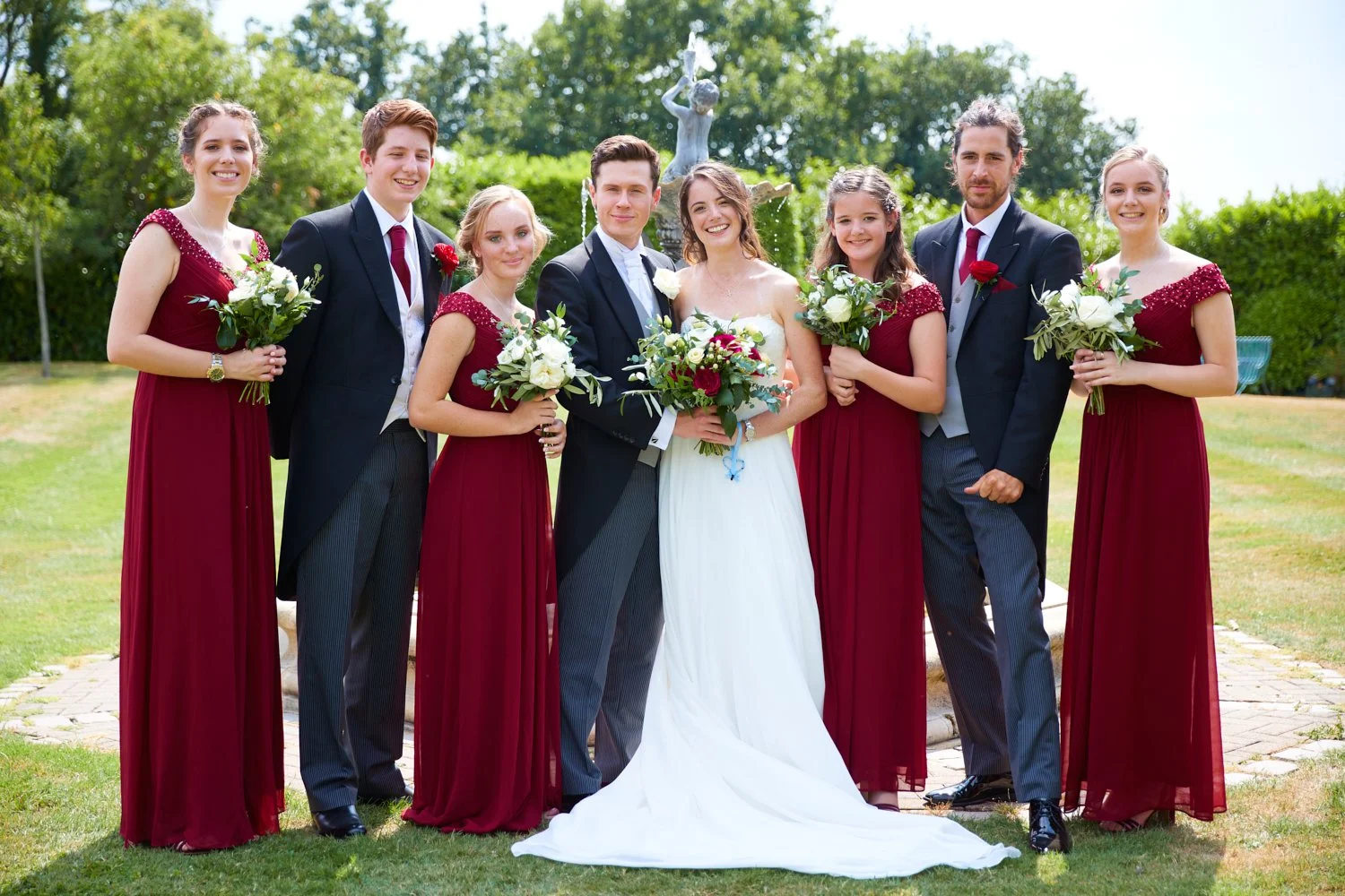 Group of wedding guests, including bride and groom, posing outdoors with bouquets of flowers, with trees and a fountain in the background.