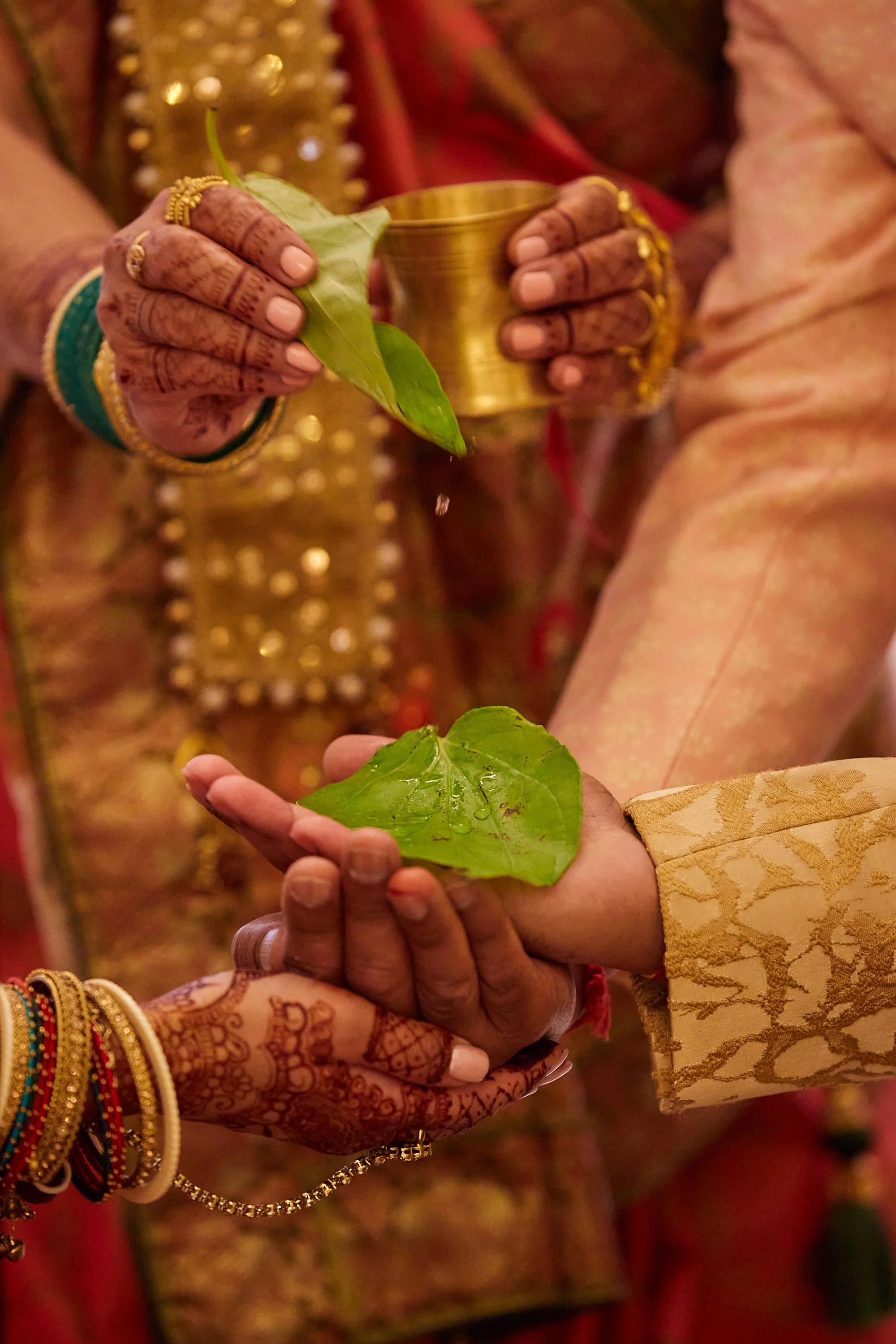 Close-up of people’s hands during a traditional ceremony, holding green leaves and a small golden container, with henna designs on the hands.
