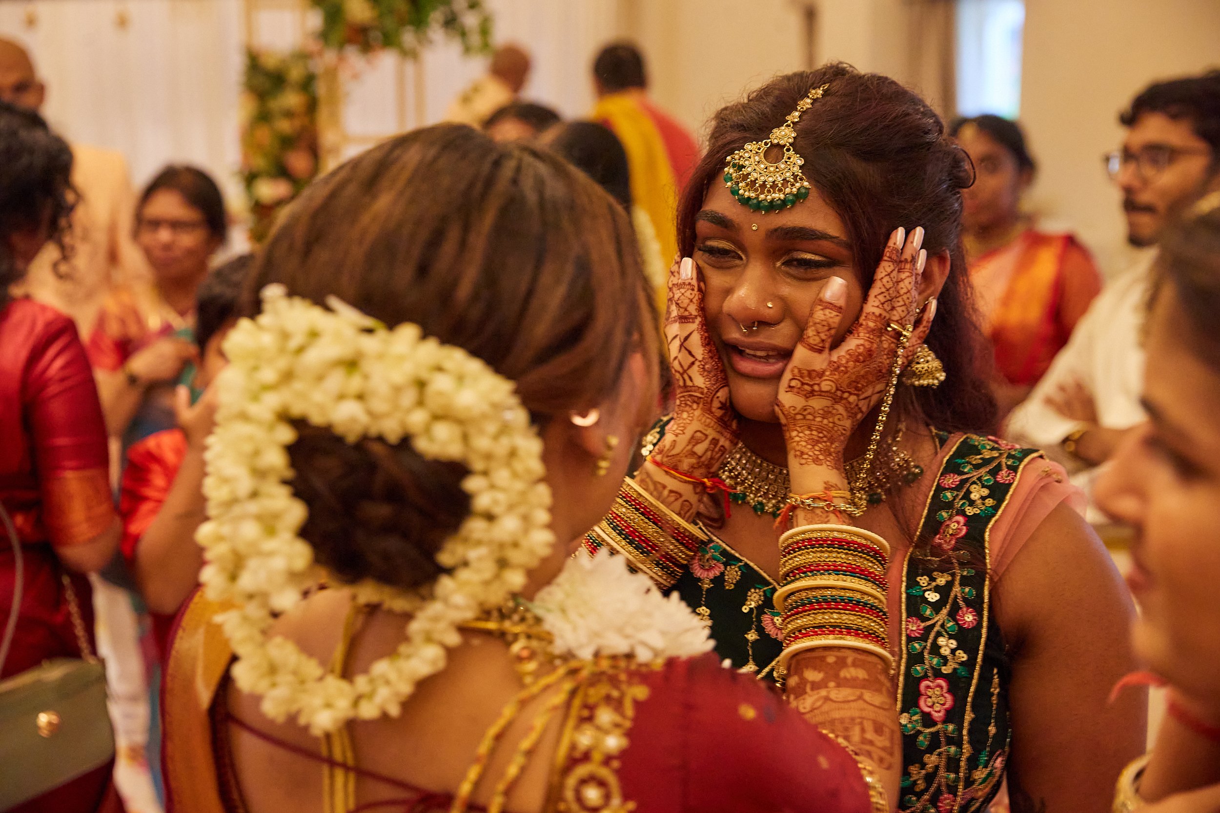 A woman dressed in traditional Indian attire is emotionally crying while someone offers comfort at a celebration, with other guests dressed in vibrant traditional clothing in the background.